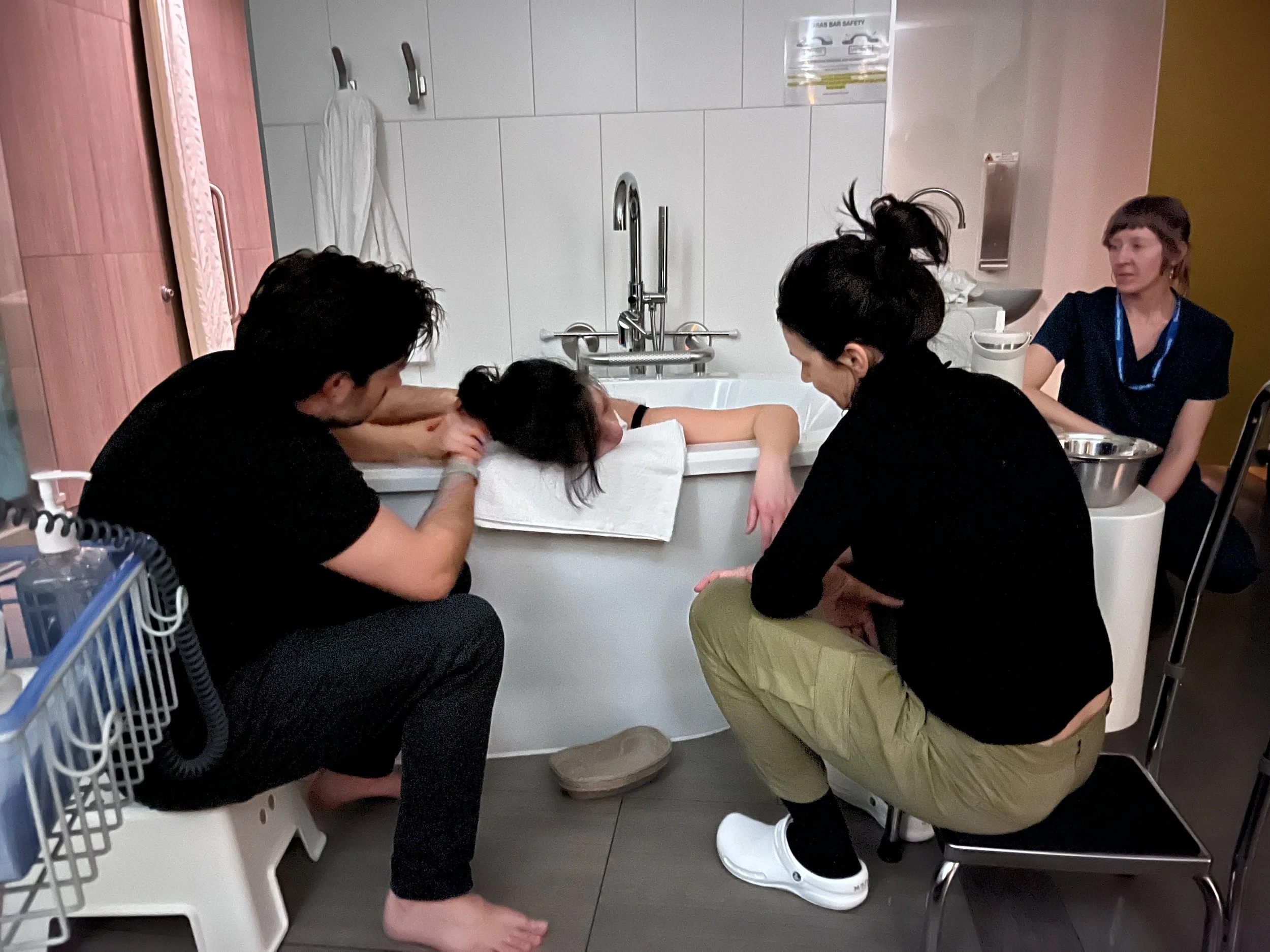 Geraldine Dempsey, Toronto-based doula, is assisting a woman in a hospital birth in a bath. A nurse with short hair and a blue lanyard is observing nearby.