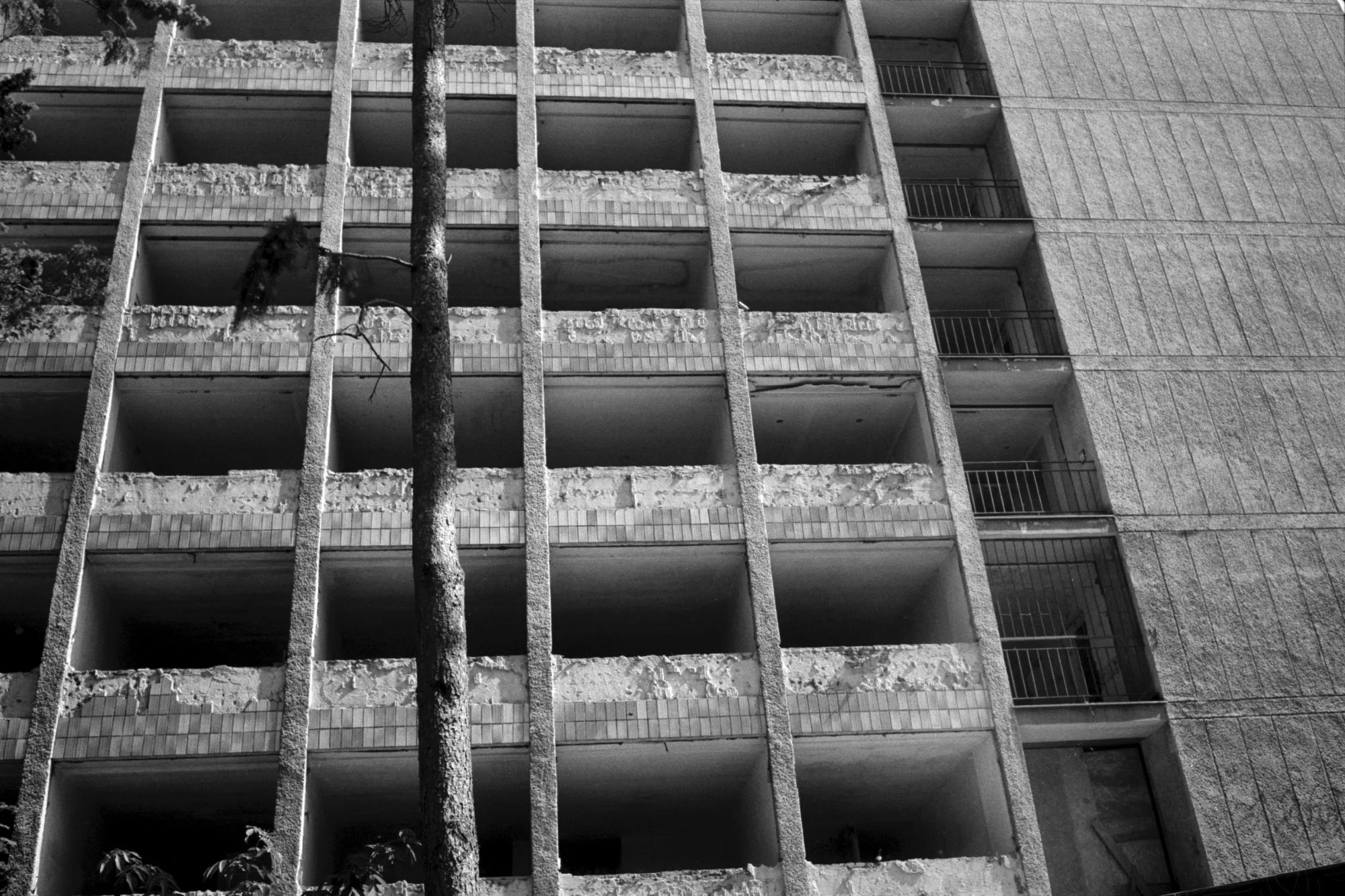 Black and white photo of an old, abandoned 5-story apartment building with exposed, damaged concrete and peeling paint. The building has empty balconies with metal railings and a tree growing in front.