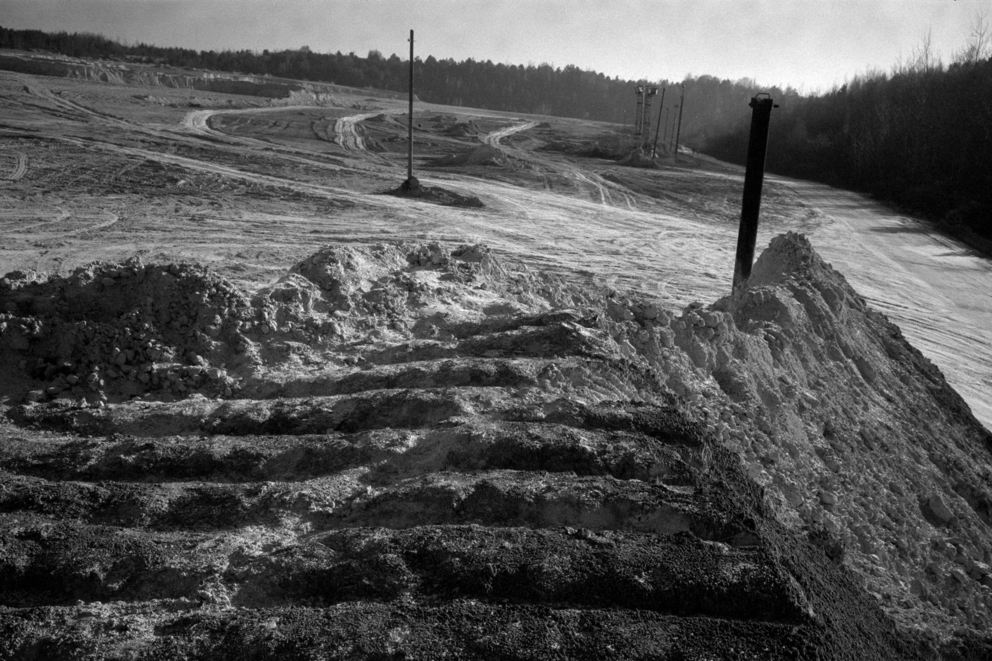 Snow-covered ski slope with mounds of snow and dark poles, with ski tracks visible and a line of trees in the background.