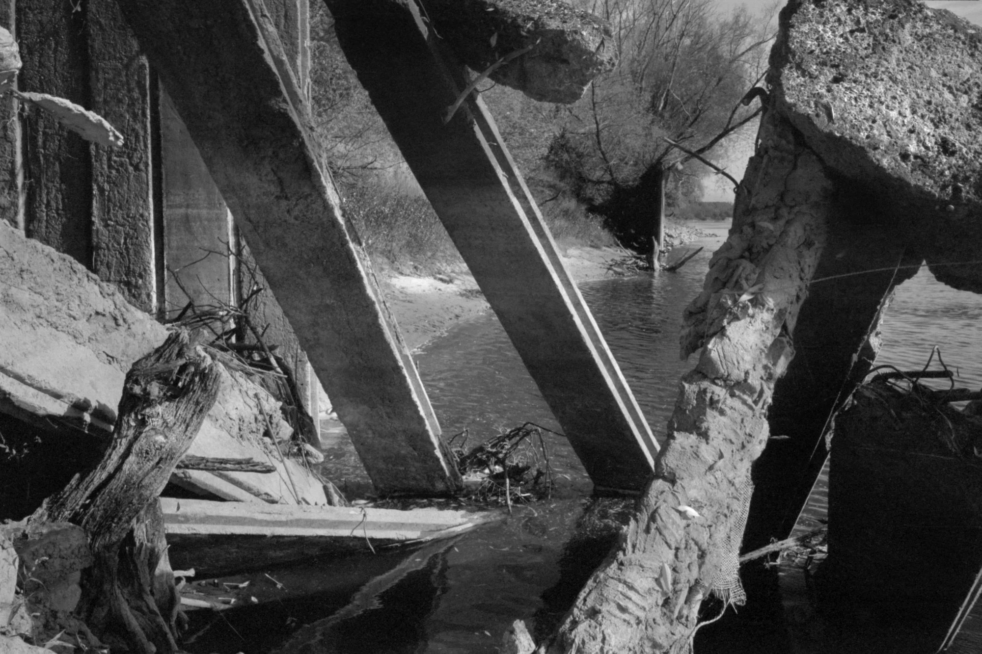 Debris and broken concrete in and around a river, part of a collapsed bridge on a winter day with leafless trees in the background.