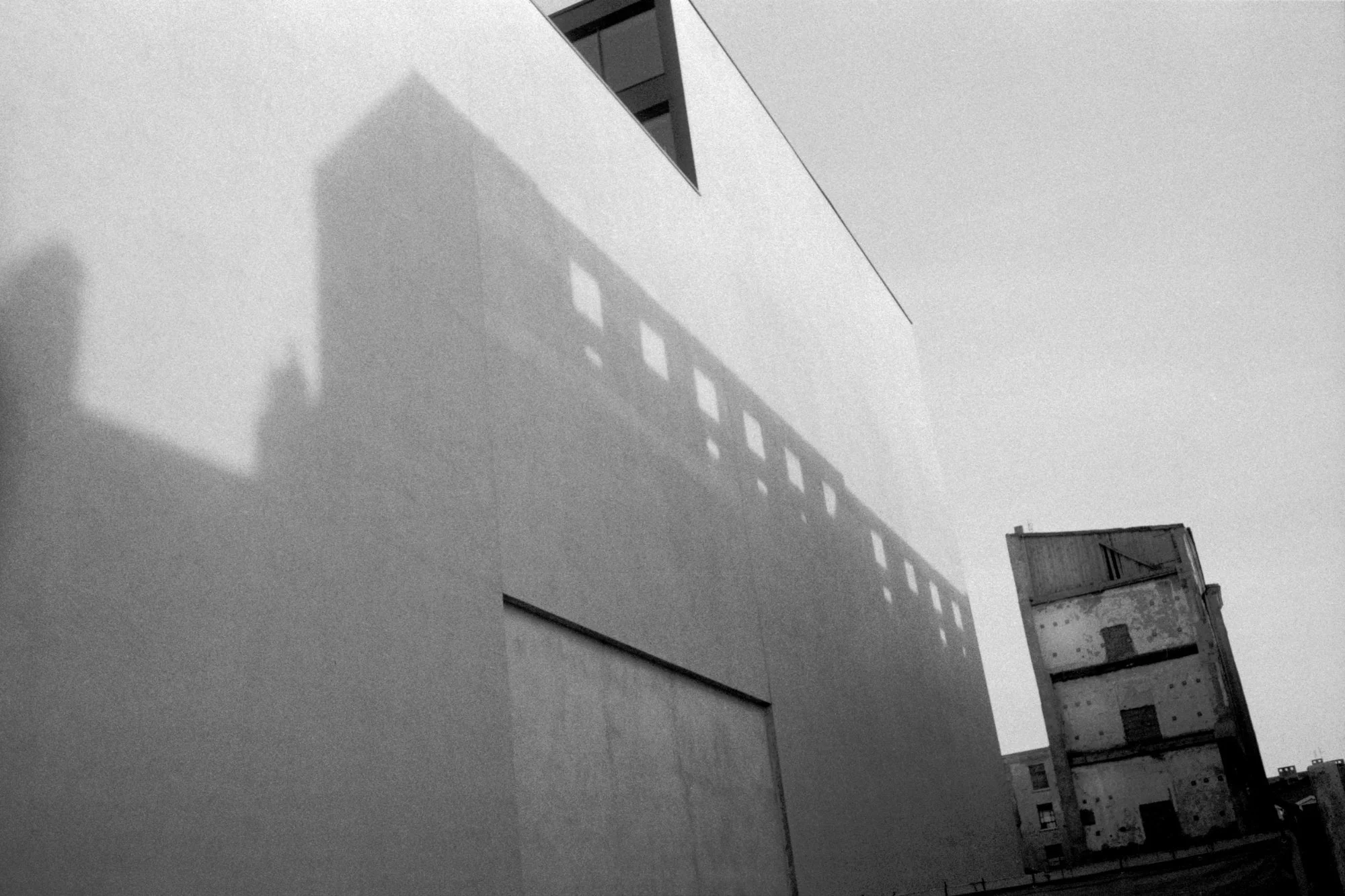Black and white photo of buildings with shadows cast on a wall, including an older, worn-down building visible in the background.
