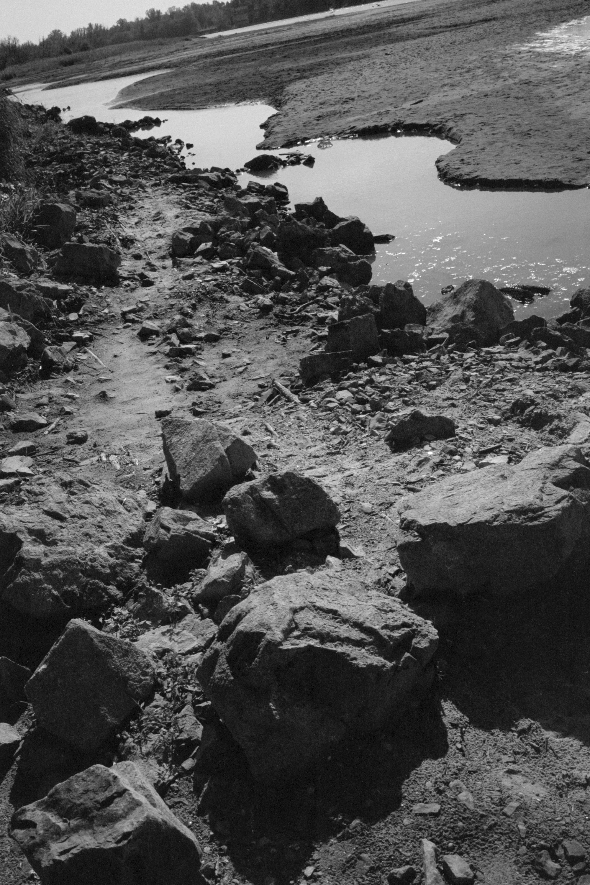 A black and white photo of a rocky dirt trail alongside a river with a gentle bend.