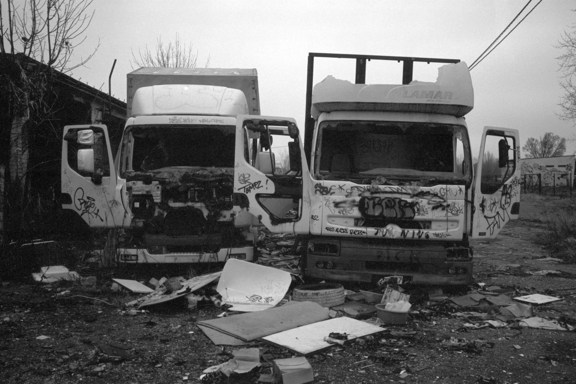 Two abandoned and heavily damaged trucks, covered in graffiti, sit amidst debris and trash in an outdoor lot, with a building and leafless trees in the background.