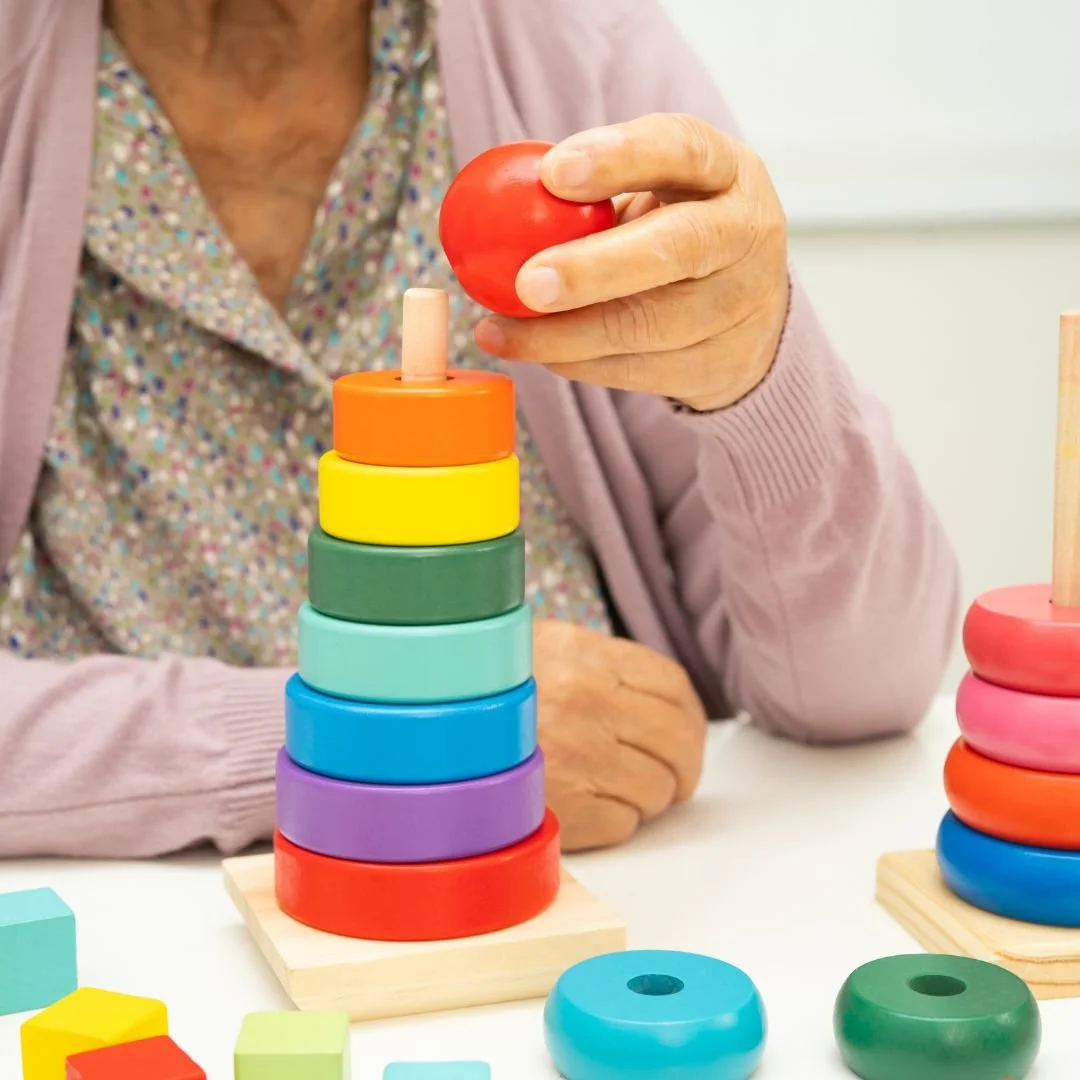 Persona anziana che impila anelli di diversi colori su un supporto, con alcuni anelli colorati sul tavolo.