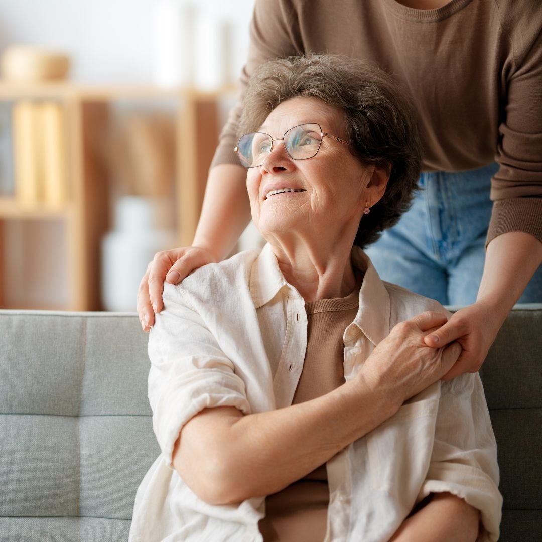Una donna anziana con occhiali sorride guardando avanti mentre una persona la abbraccia da dietro in un ambiente domestico.