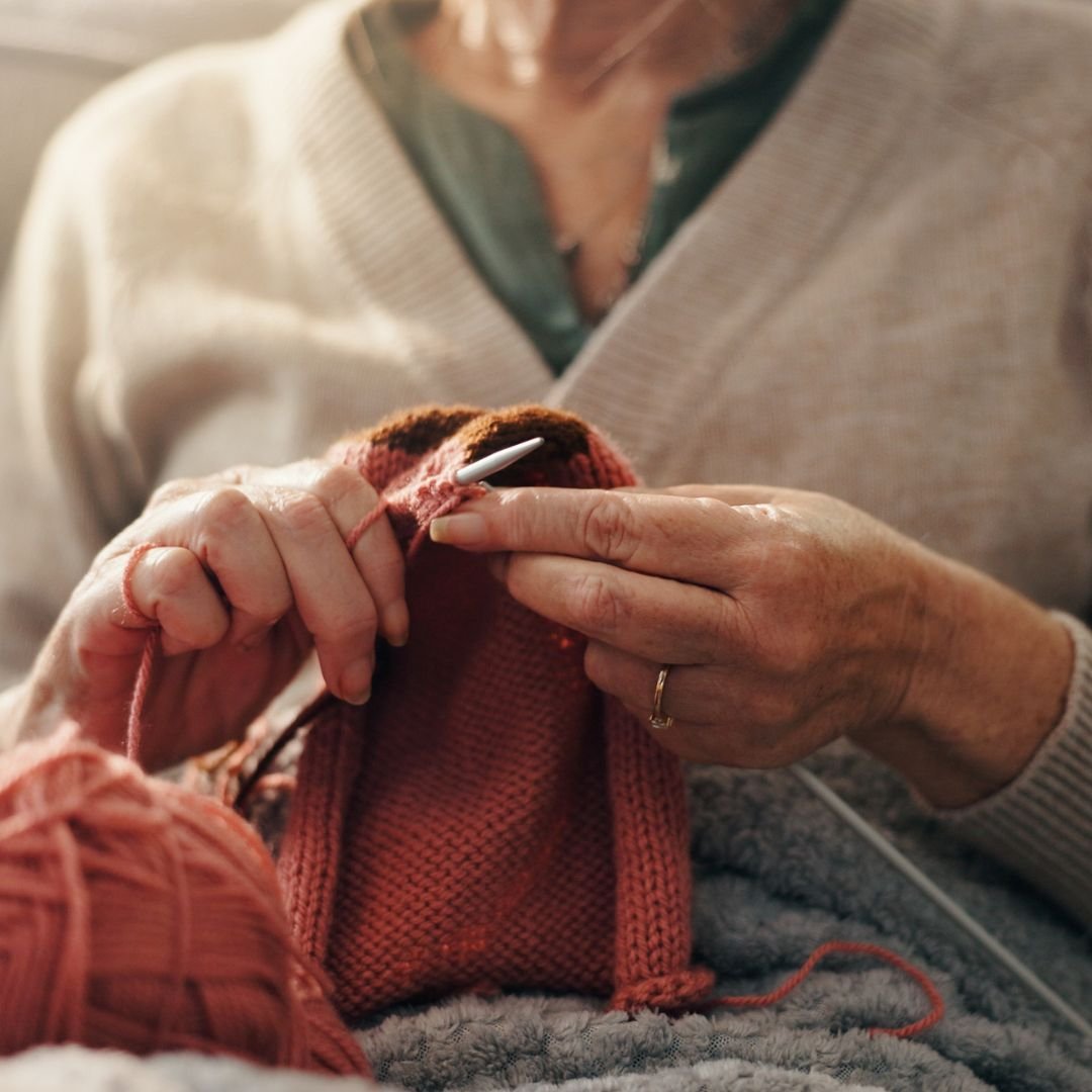 Una persona anziana che cuce un maglione di lana rosso, utilizzando un ago. Le mani sono visibilmente anziane, con evidenti segni di invecchiamento.