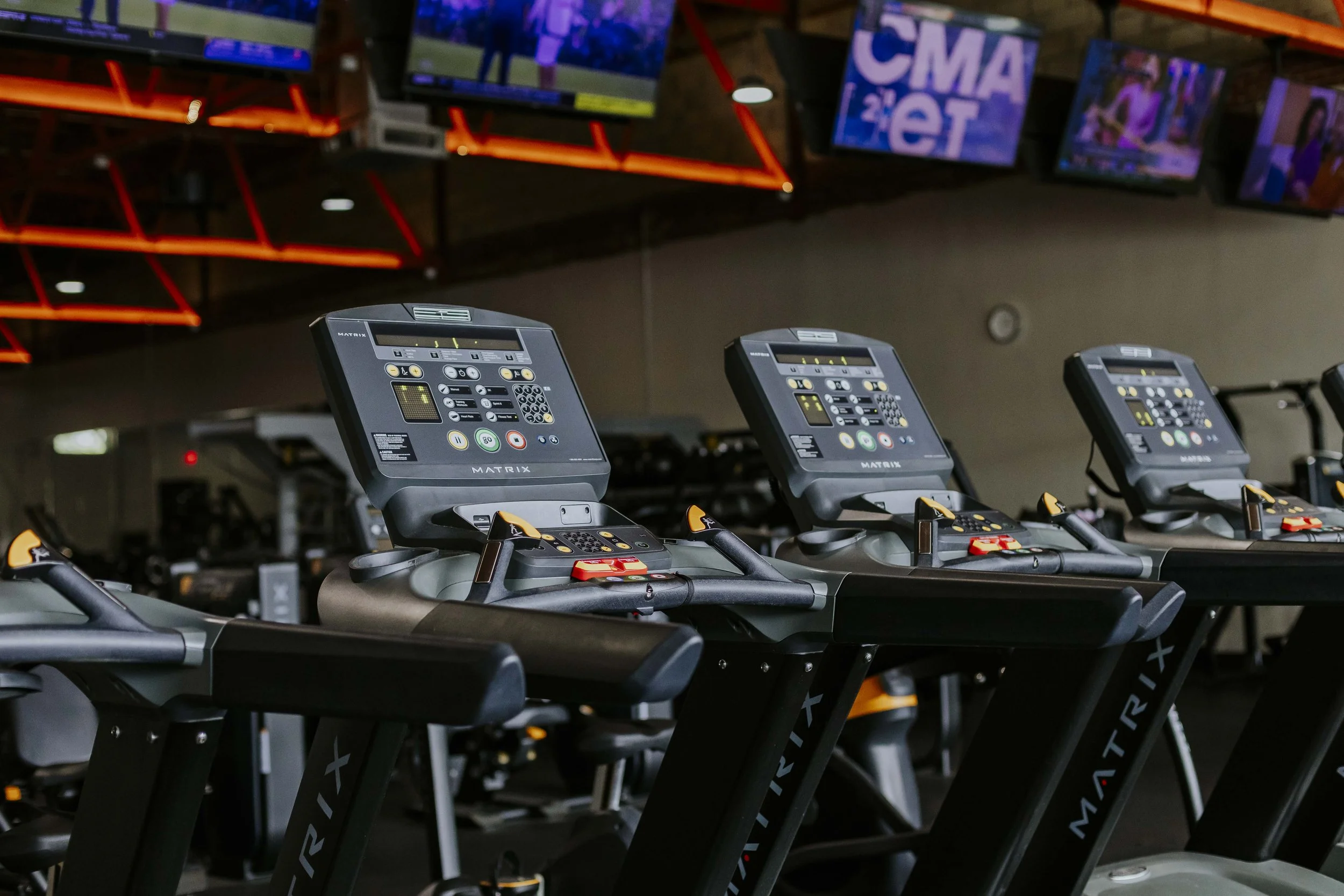 Indoor gym with three treadmills in the foreground and multiple TV screens mounted on the wall in the background displaying various images.