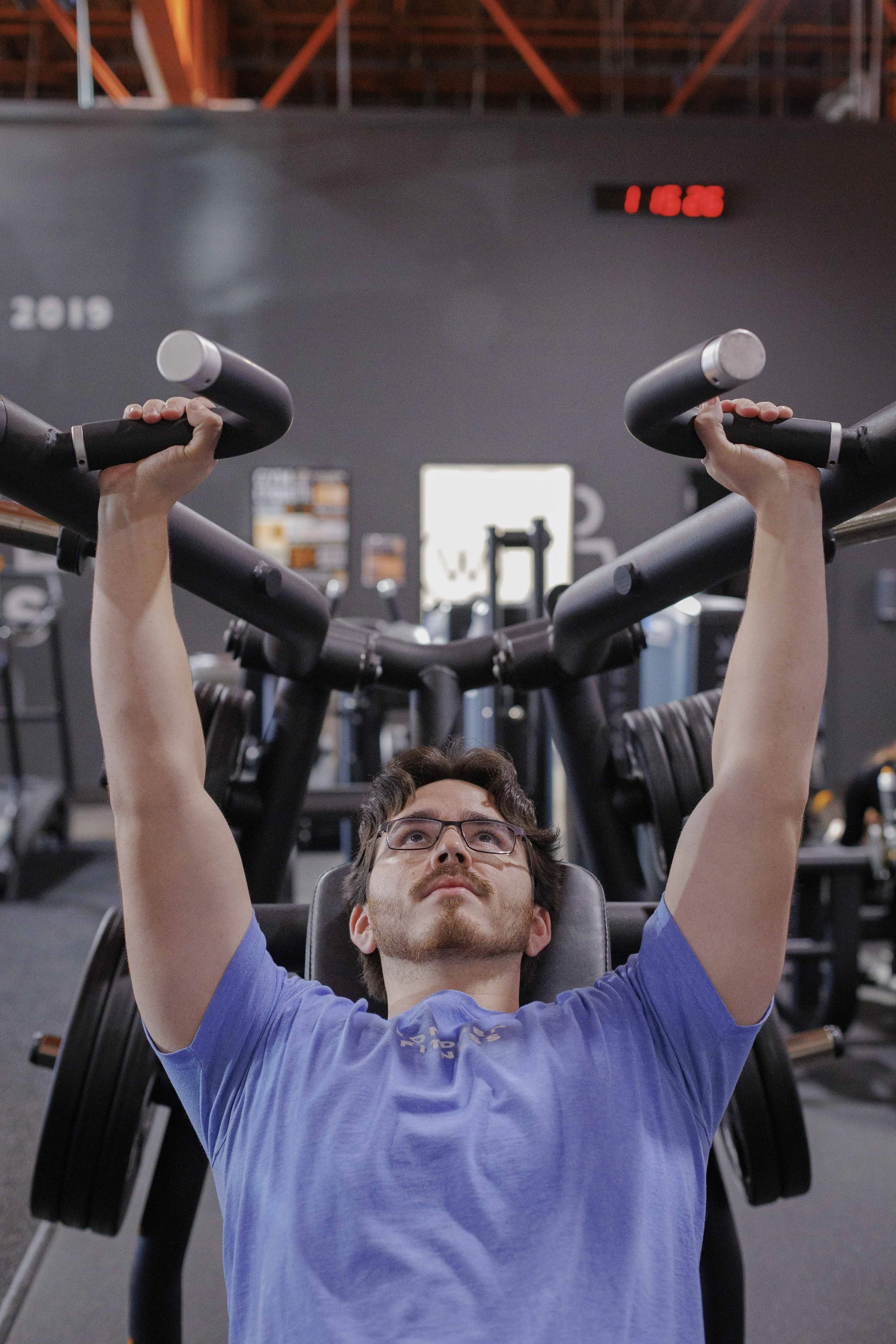 A man with glasses in a blue t-shirt lying on a weight bench, lifting dumbbells in a gym with gym equipment and a dark wall in the background.