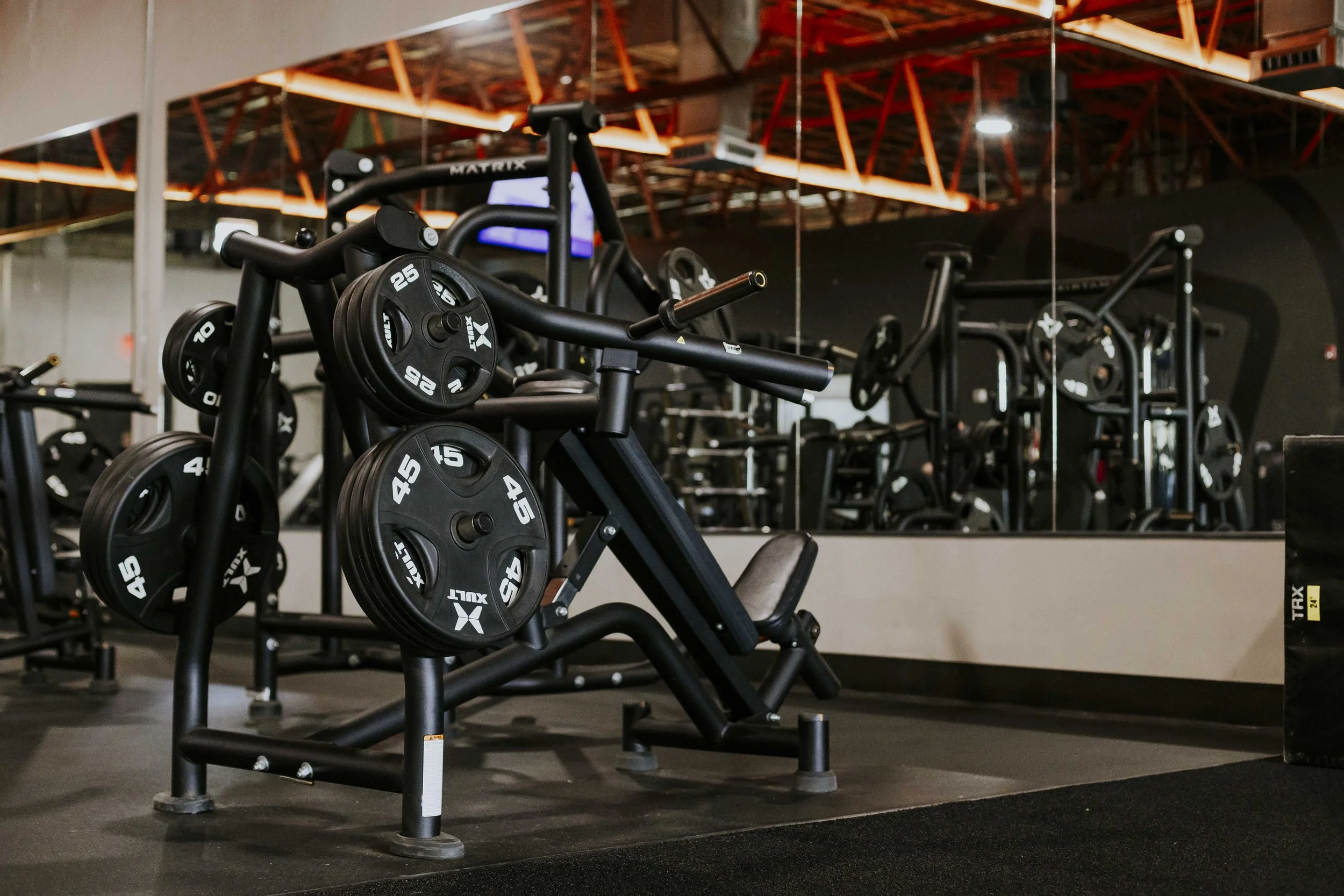 A gym with weightlifting equipment, including black weight plates with white numbers, placed on a seated leg press machine. The background features a large mirror reflecting other exercise machines and orange ceiling lighting.