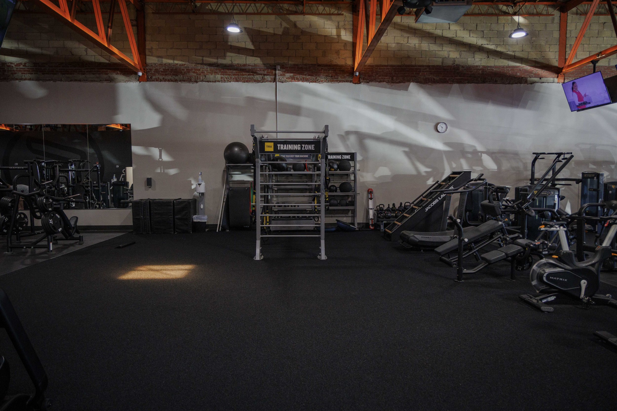 An empty gym with workout equipment including weights, exercise machines, and a training zone area with shelves of medicine balls and kettlebells. The gym has exposed brick and industrial-style lighting, with a wall clock and two wall-mounted TVs.