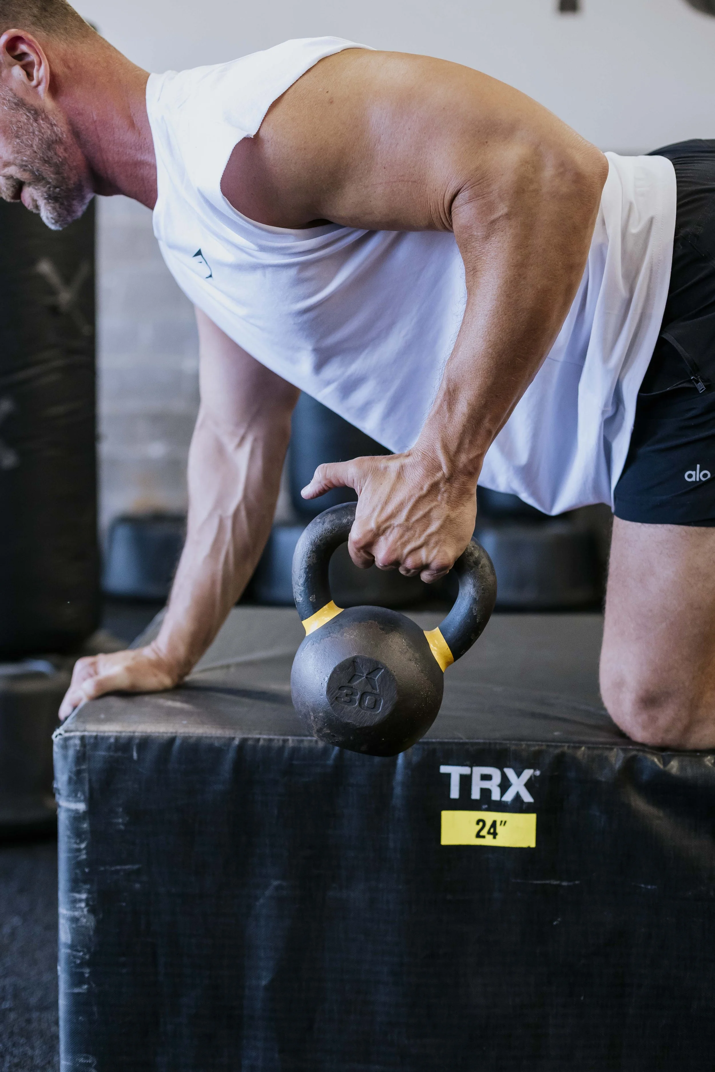 A man with gray hair and beard performing a kettlebell exercise on a black platform, wearing a white sleeveless shirt and black shorts inside a gym.