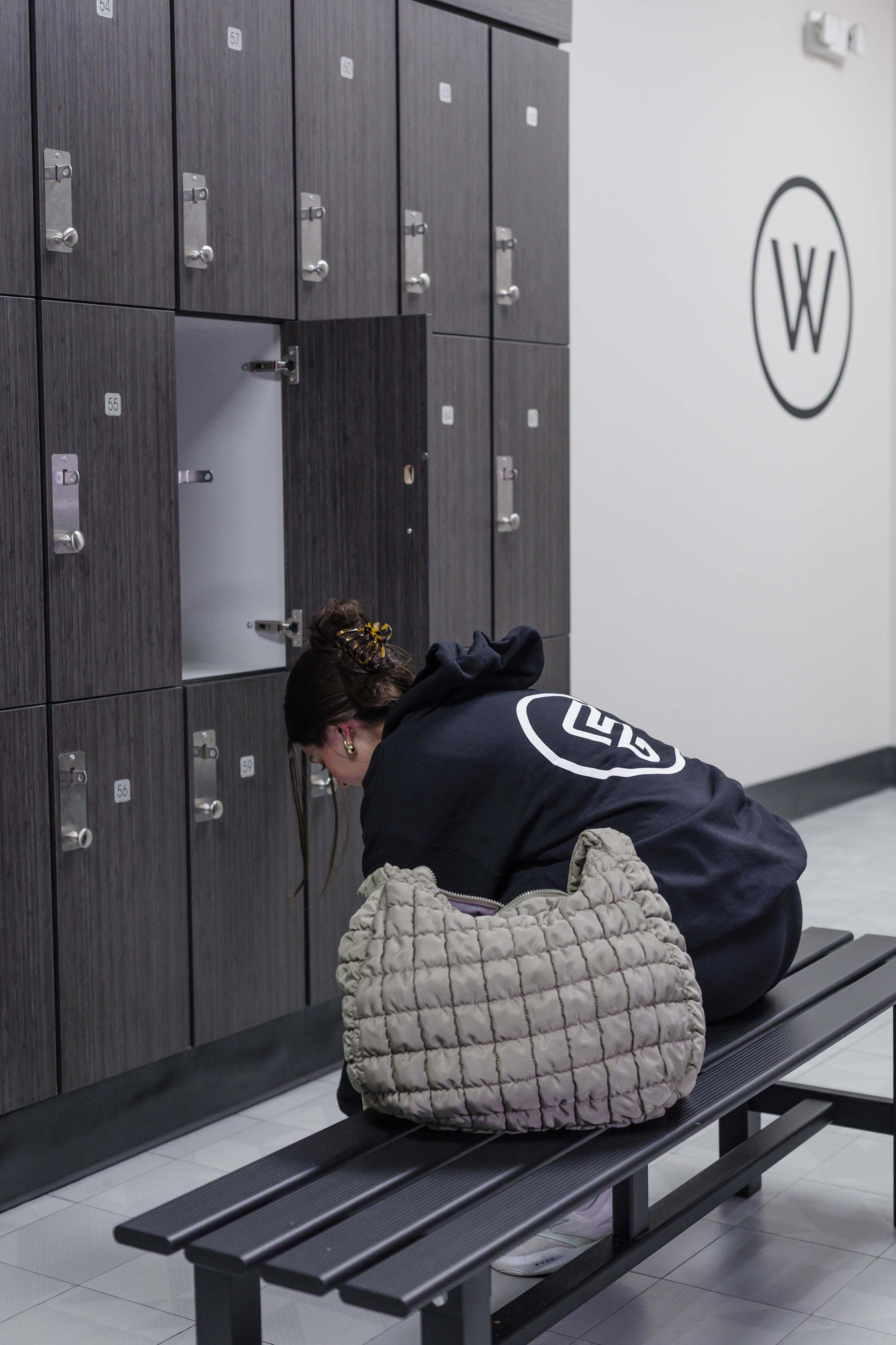 A woman with dark hair tied in a bun, wearing a black hoodie with a logo, is sitting on a black bench at an airport or train station. She is looking down at her phone or something in her lap. Next to her, a beige quilted jacket is draped over a chair
