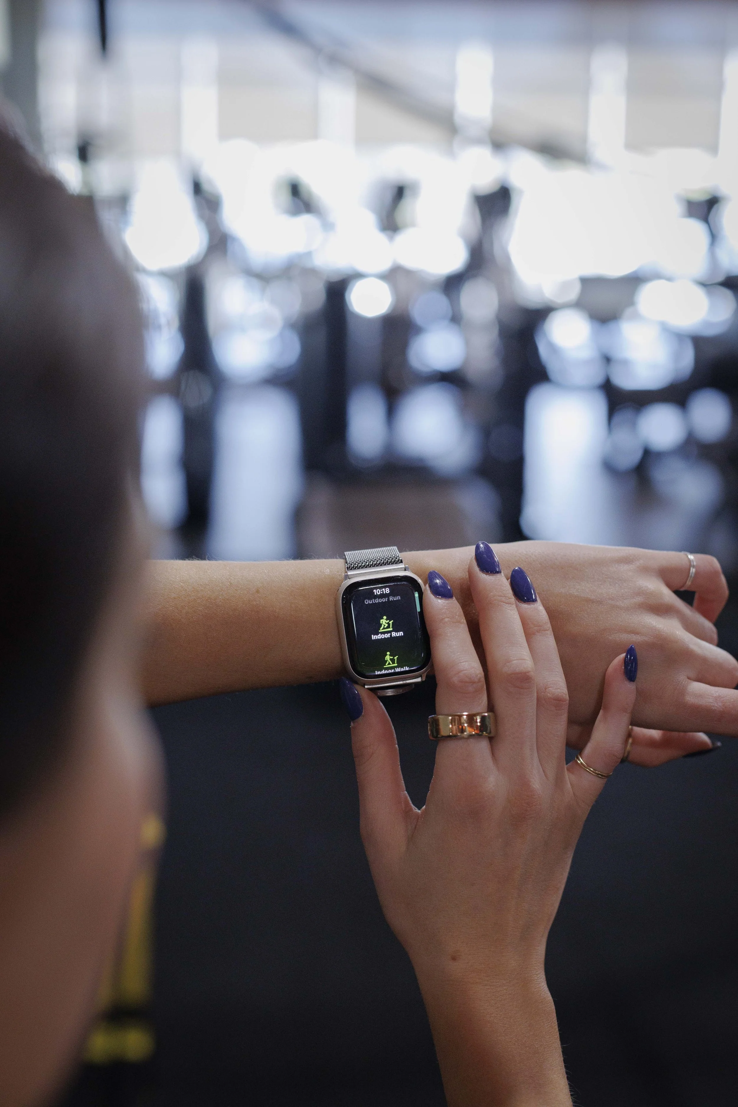 Close-up of a woman's left wrist with a smartwatch displaying exercise options, wearing rings and painted purple nails, with a blurred background of indoor space and other people.
