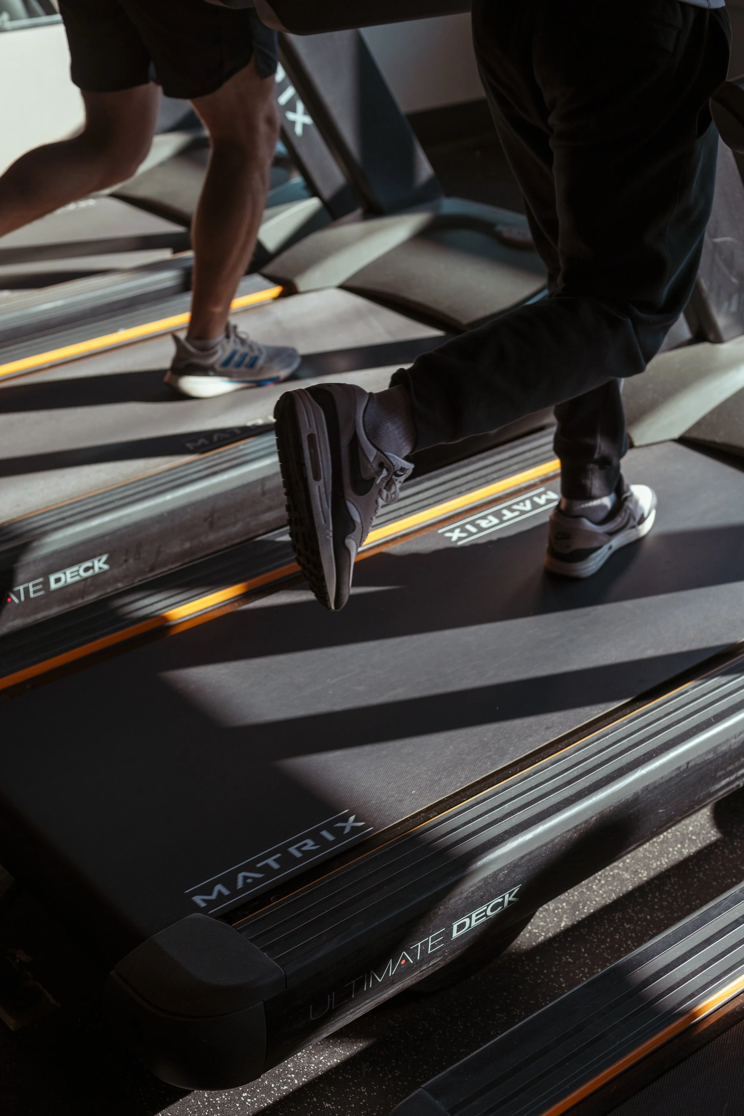 Close-up of two people using treadmill machines labeled 'Ultimate Deck' in a gym.