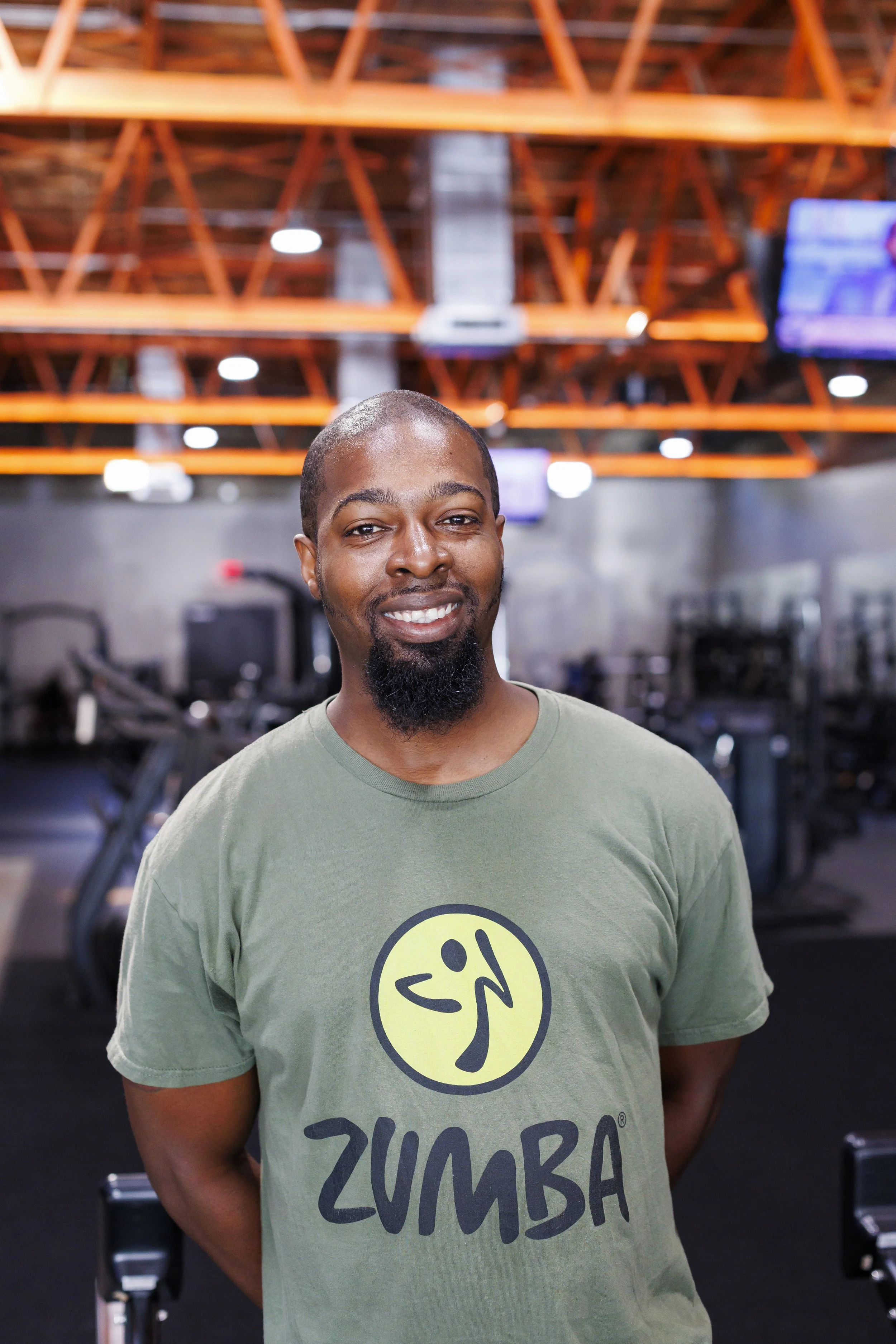 Smiling man with short hair and beard in a gym, wearing a green T-shirt with 'Zumba' logo, with gym equipment and a television screen in the background.