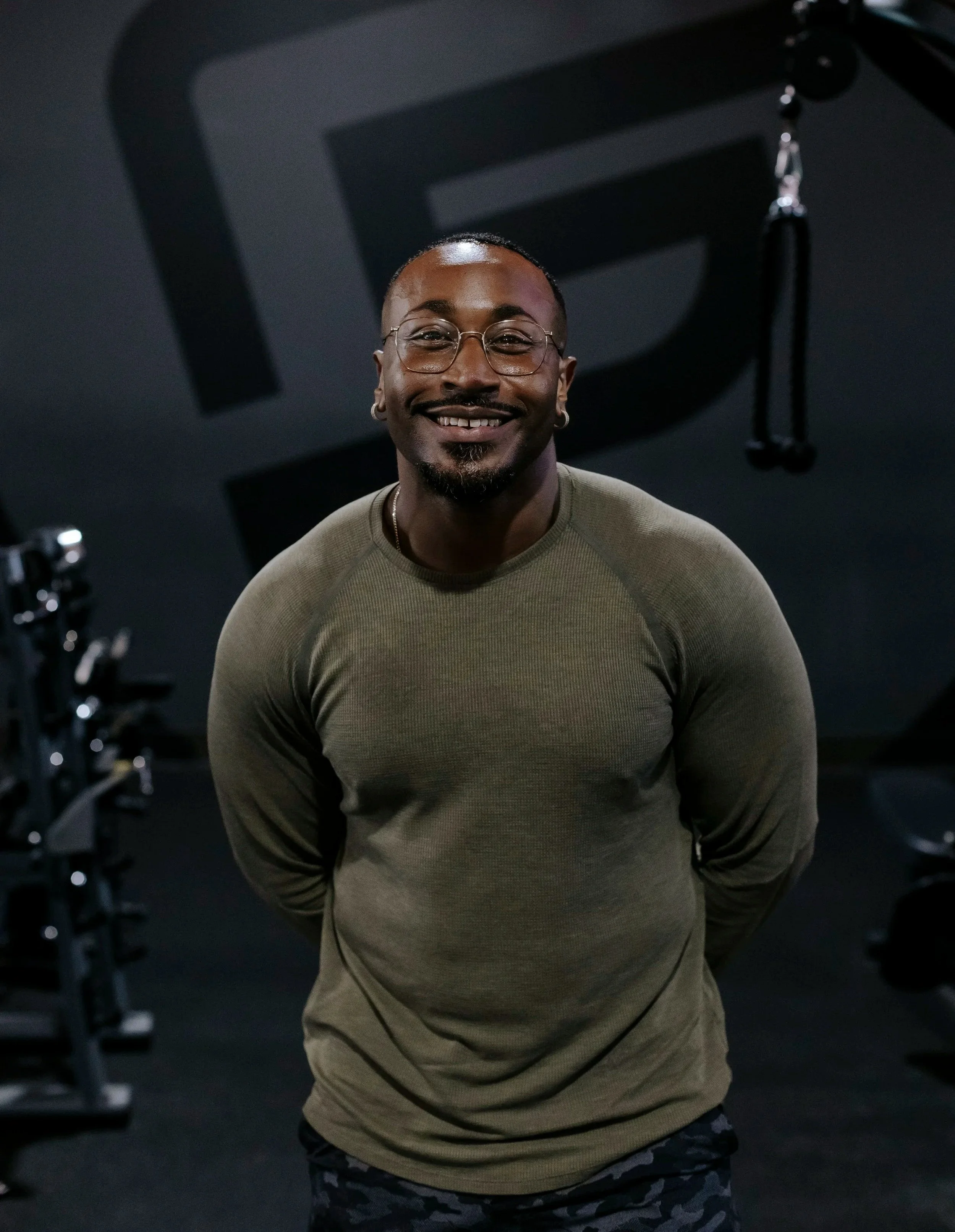 A man with glasses and earrings smiling in a gym with workout equipment and a black ceiling with the letter 'D' in the background.