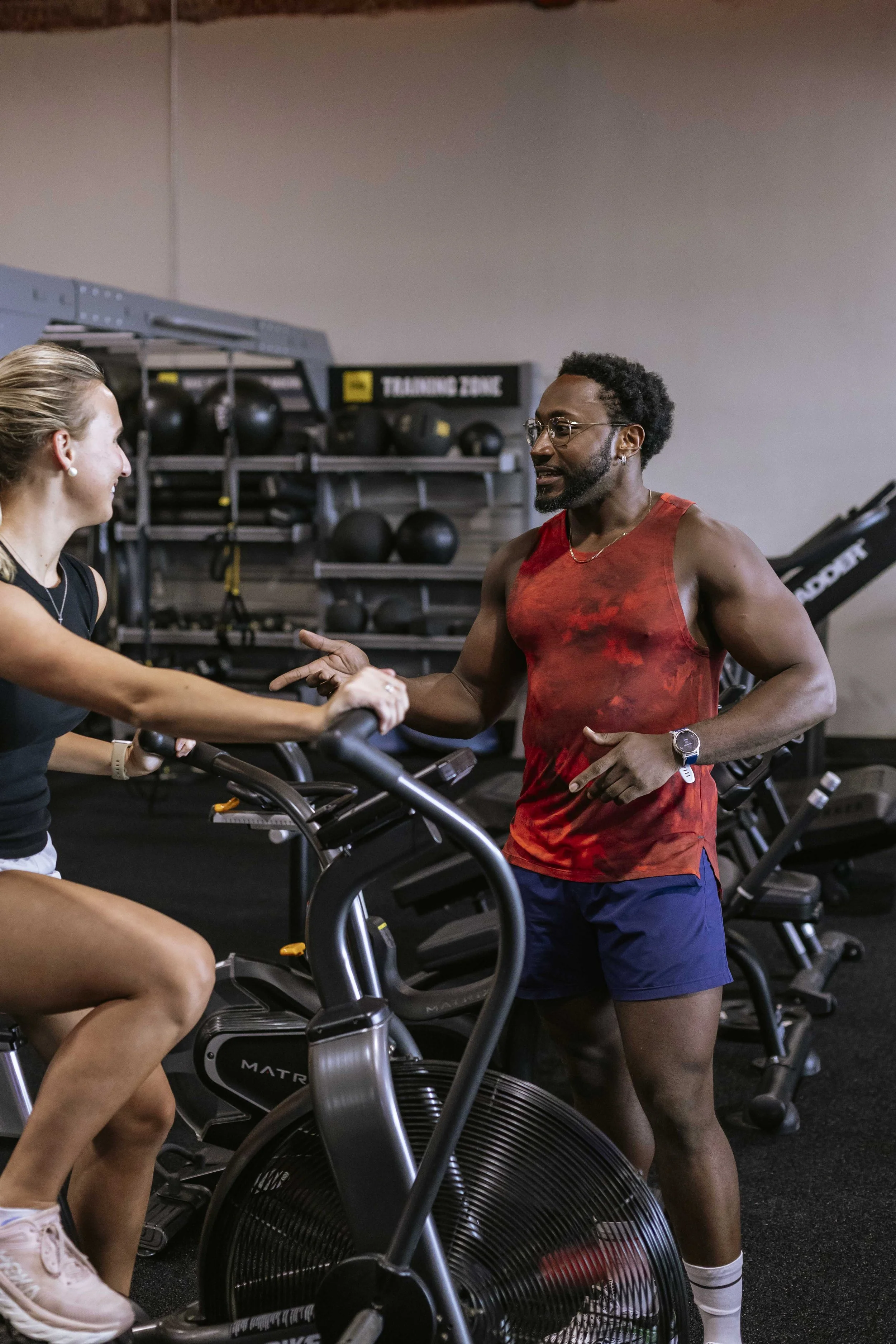 A man and woman in a gym, shaking hands. The woman is on an exercise bike, wearing a black tank top and white sneakers. The man is standing next to her, wearing a red sleeveless shirt, navy shorts, white socks, and glasses. They are indoors, with gym