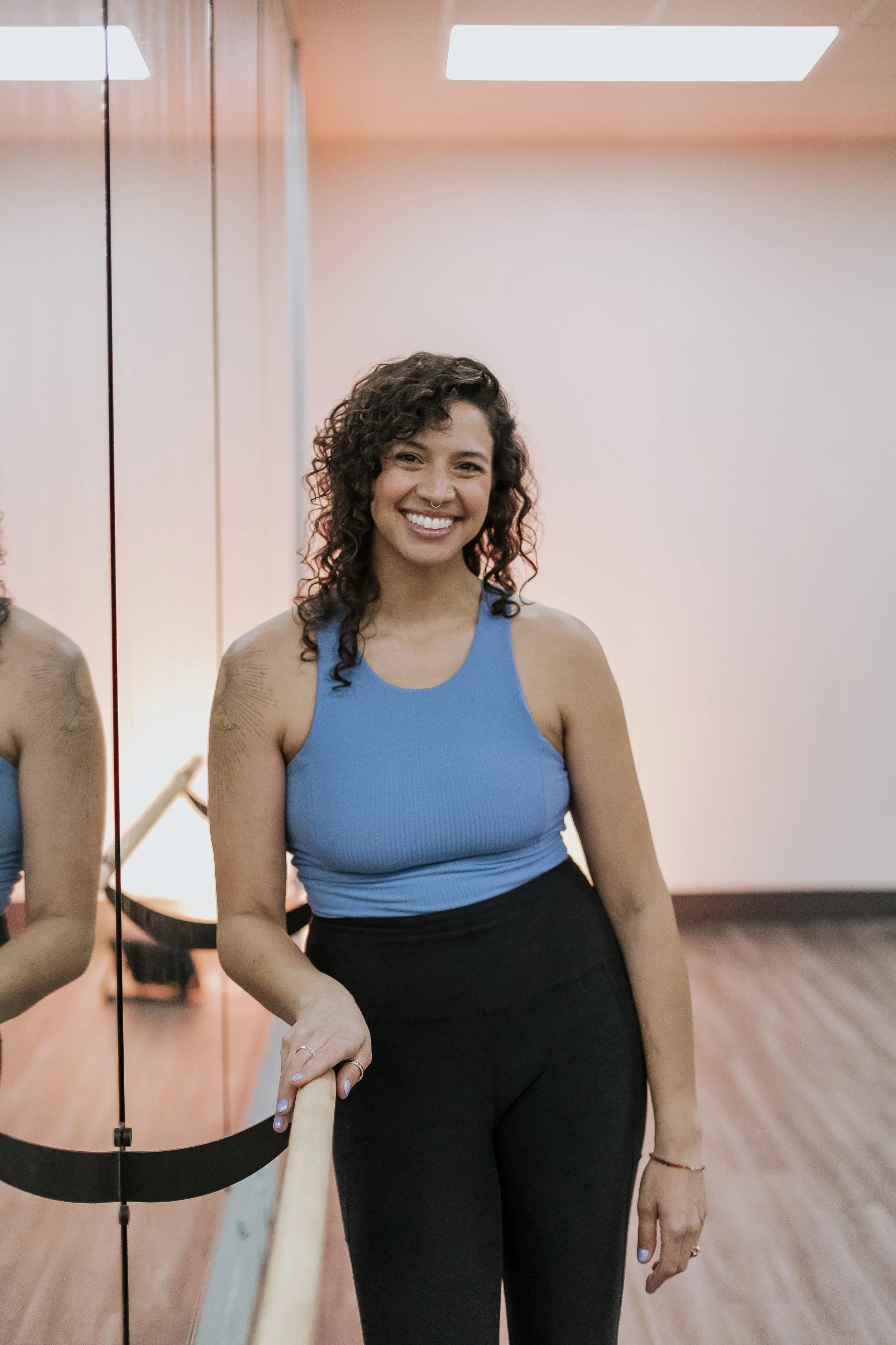 A woman with curly hair smiling, wearing a blue sleeveless top and black pants, standing in front of a mirror in a dance studio.