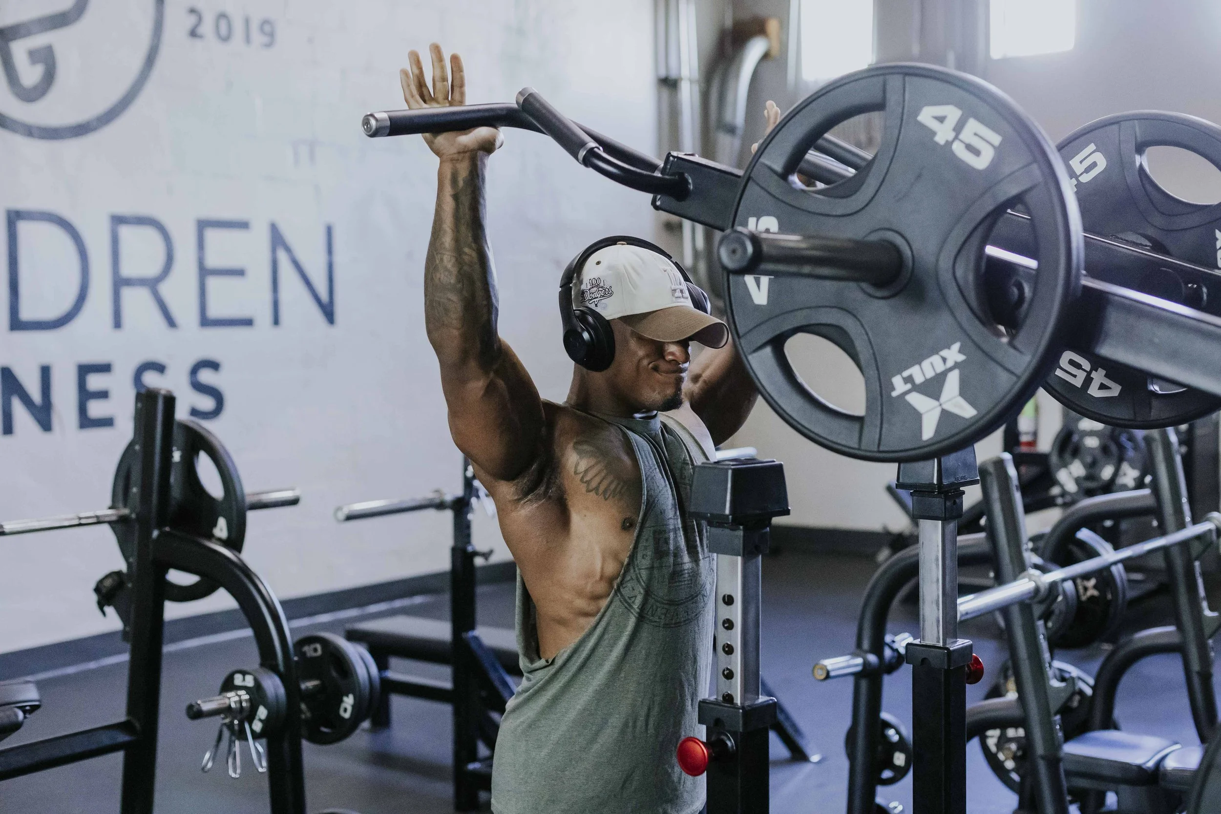 Man lifting a barbell in a gym, wearing headphones, a cap, and tank top.