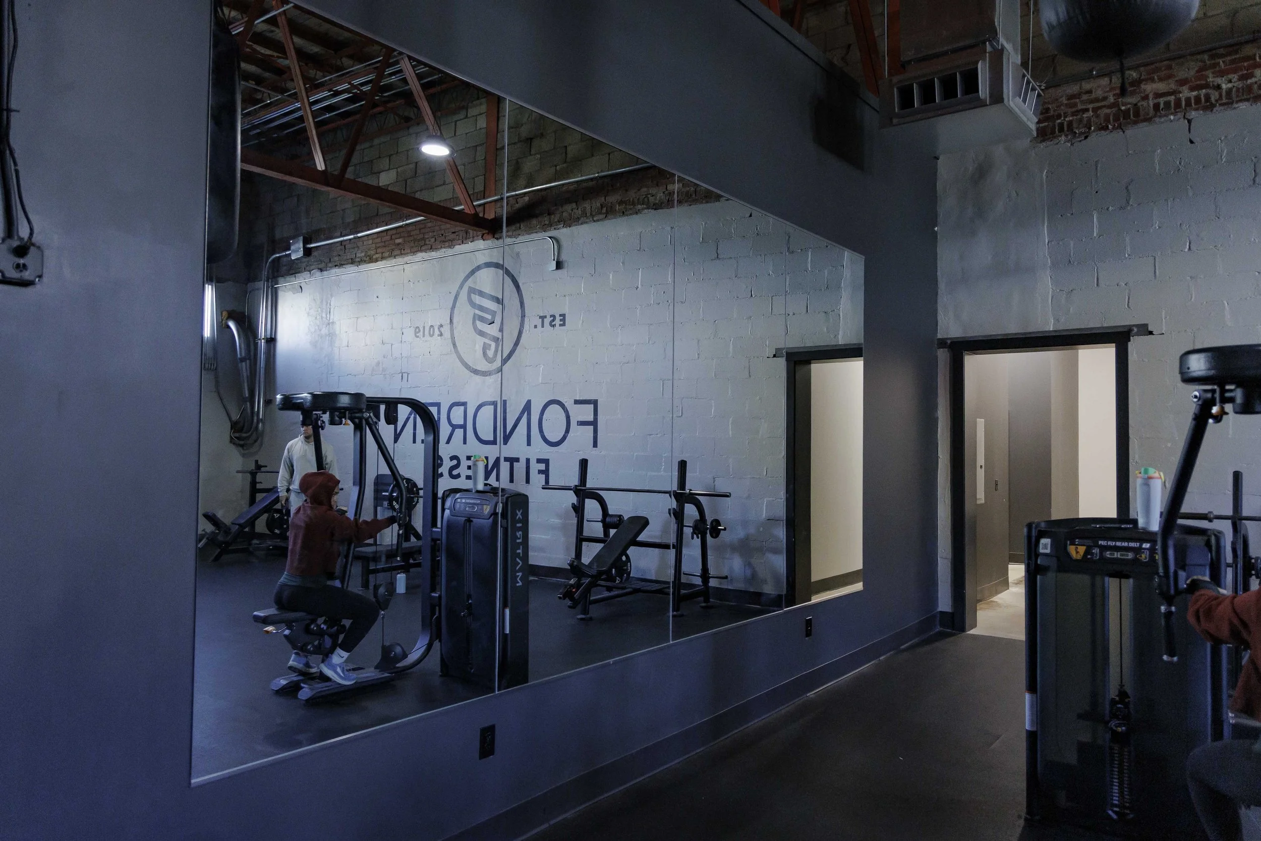 Inside a gym with exercise equipment and a mirror on the wall reflecting a person exercising. Exposed brick and industrial ceiling elements are visible.