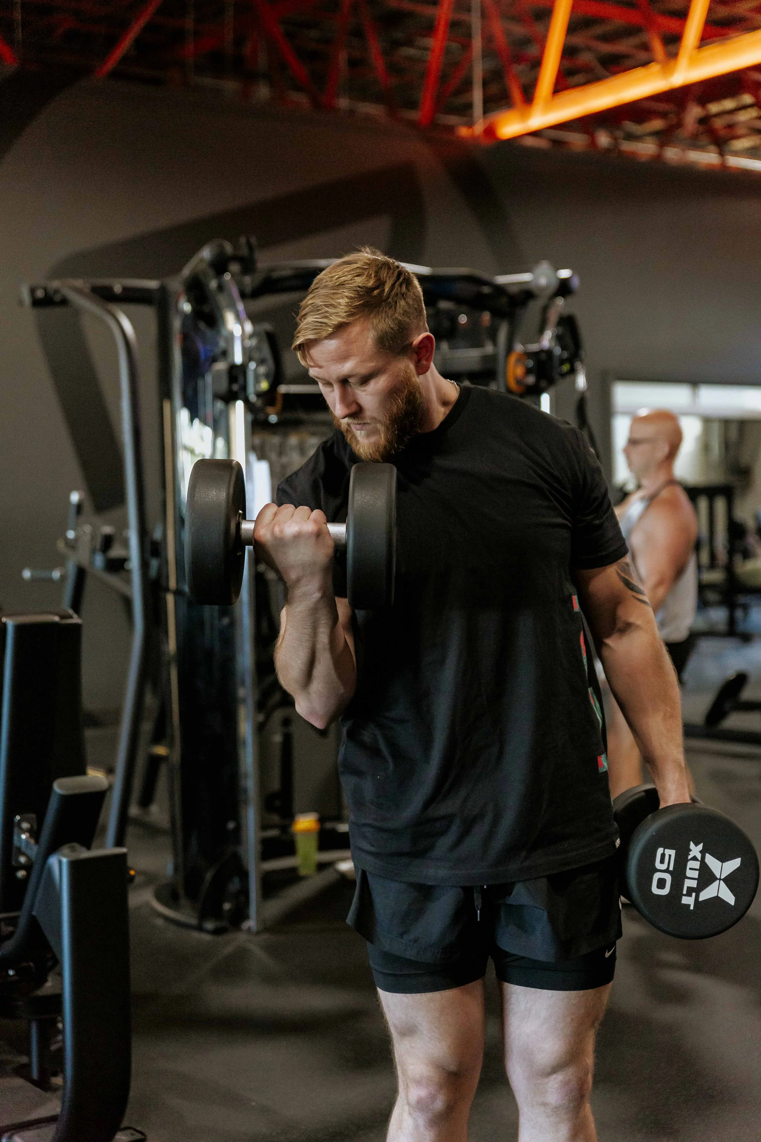A man with a beard doing bicep curls with dumbbells in a gym, with a woman in the background also lifting weights.
