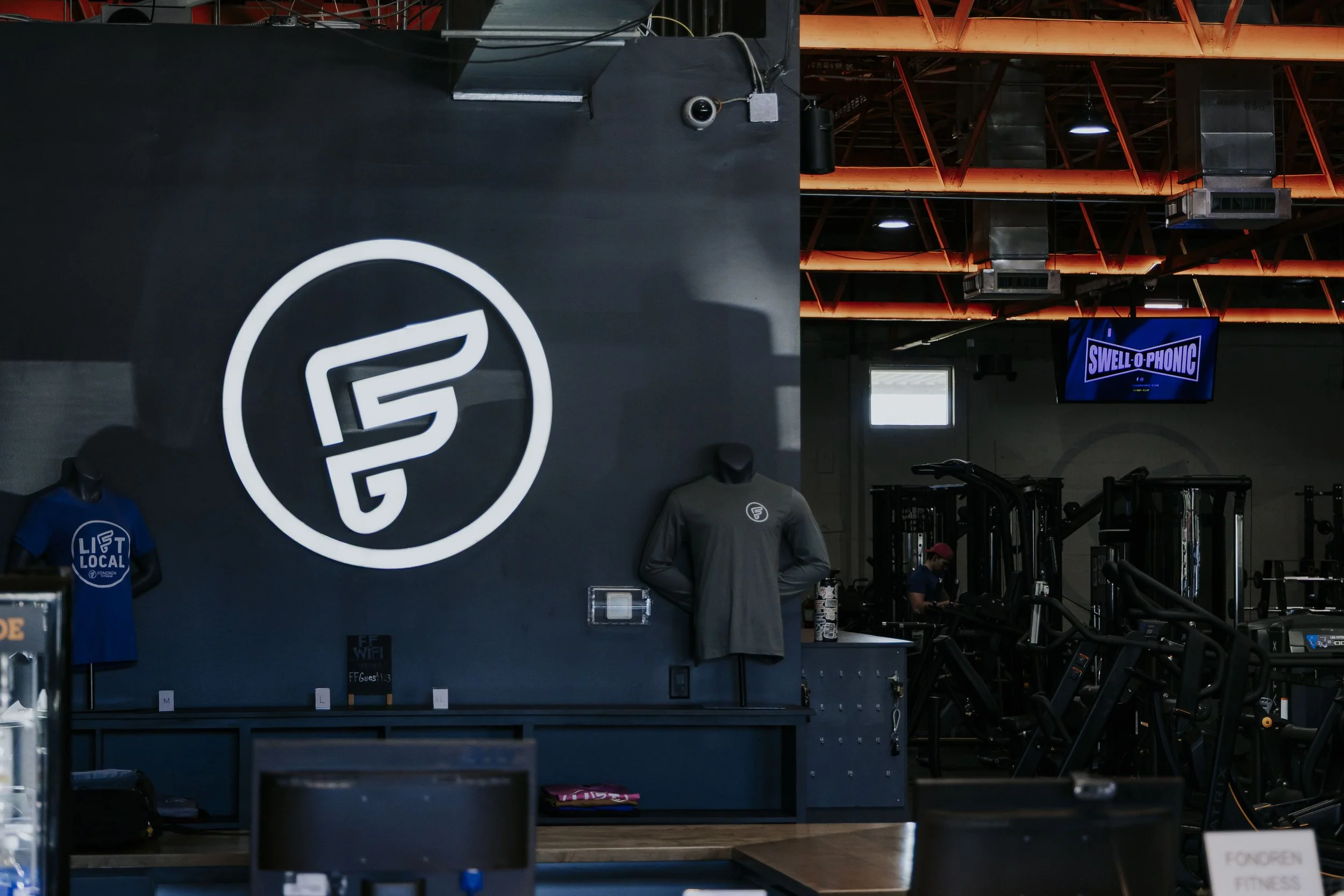 Interior of a gym with gym equipment, mannequins wearing workout shirts, and a television screen displaying the text "SWELL-O-PHONIC". The gym has black walls and orange ceiling beams.