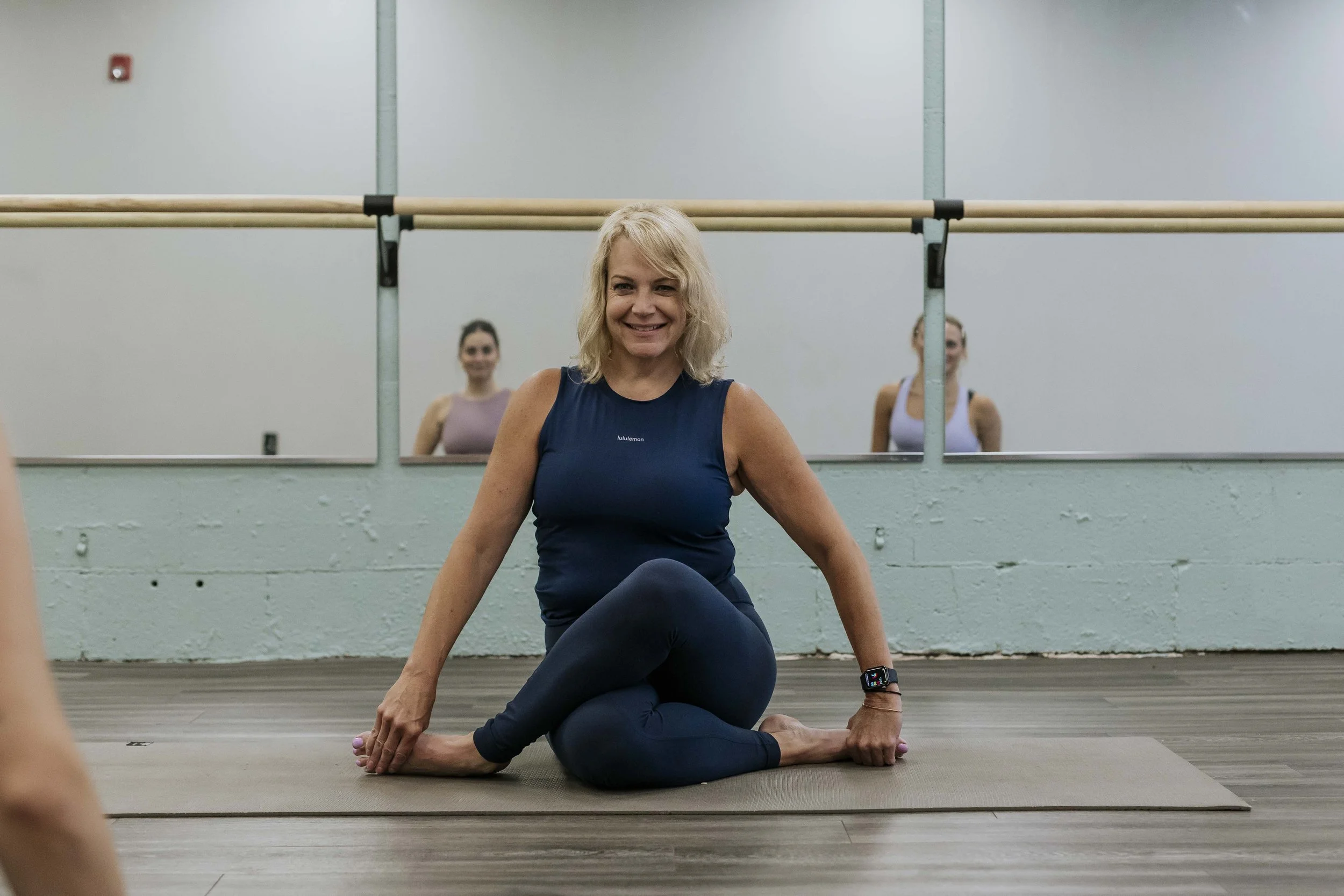 A woman in a blue athletic outfit practicing yoga indoors, sitting on a mat with her legs crossed and smiling. There are two other women in the background also doing yoga, reflected in a mirror.