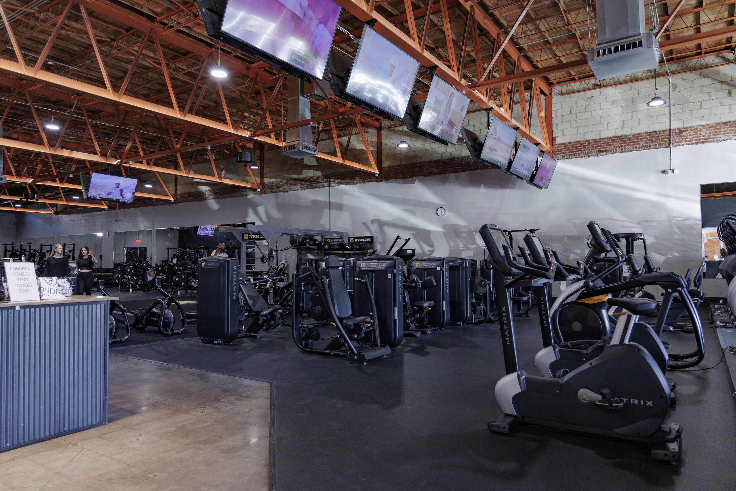 A fitness gym with exercise machines, including ellipticals and treadmills, under orange metal ceiling support beams, with a white brick wall and multiple wall-mounted televisions.