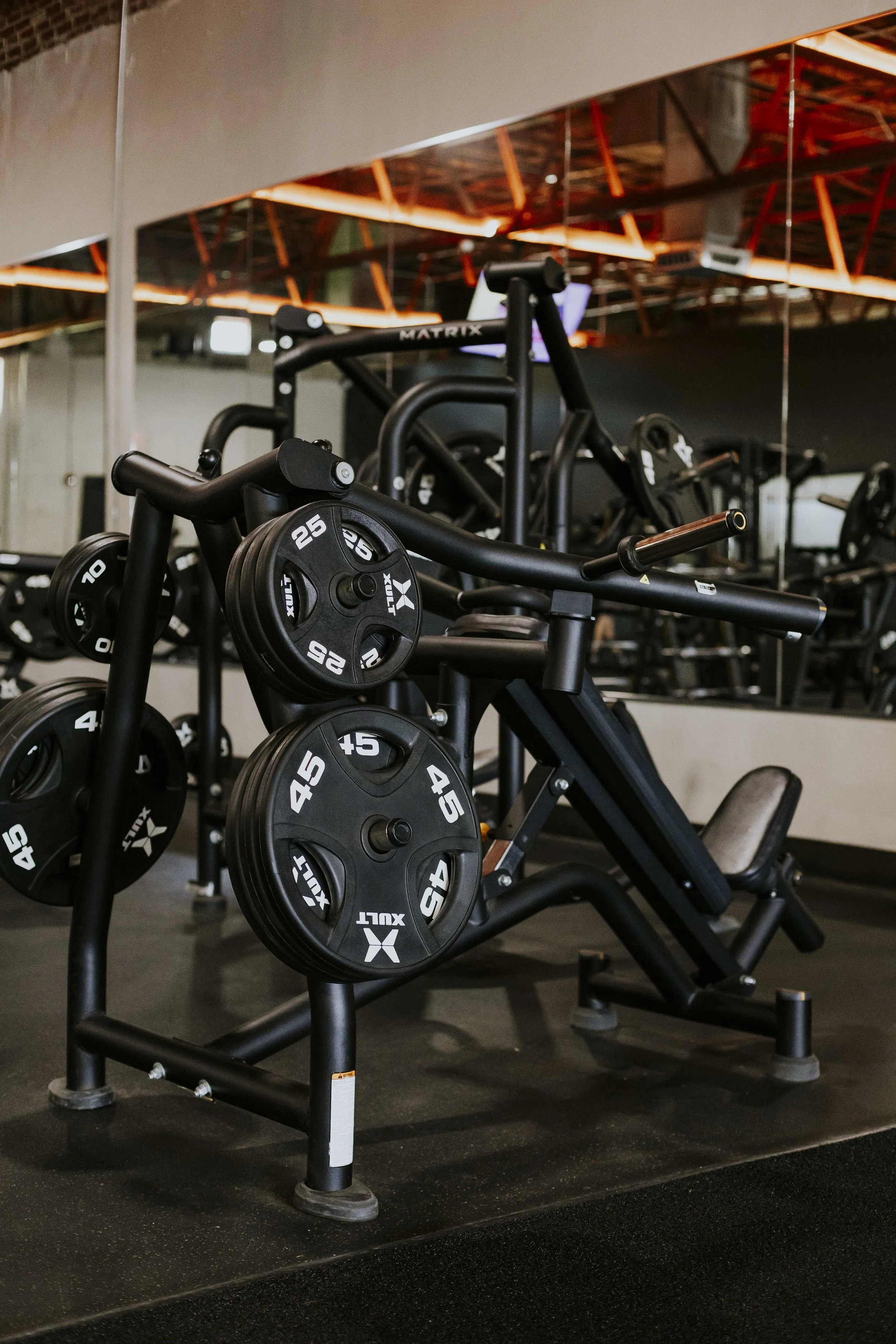 Indoor gym with black weightlifting equipment including plates and benches, illuminated by orange lighting reflected in a large mirror.