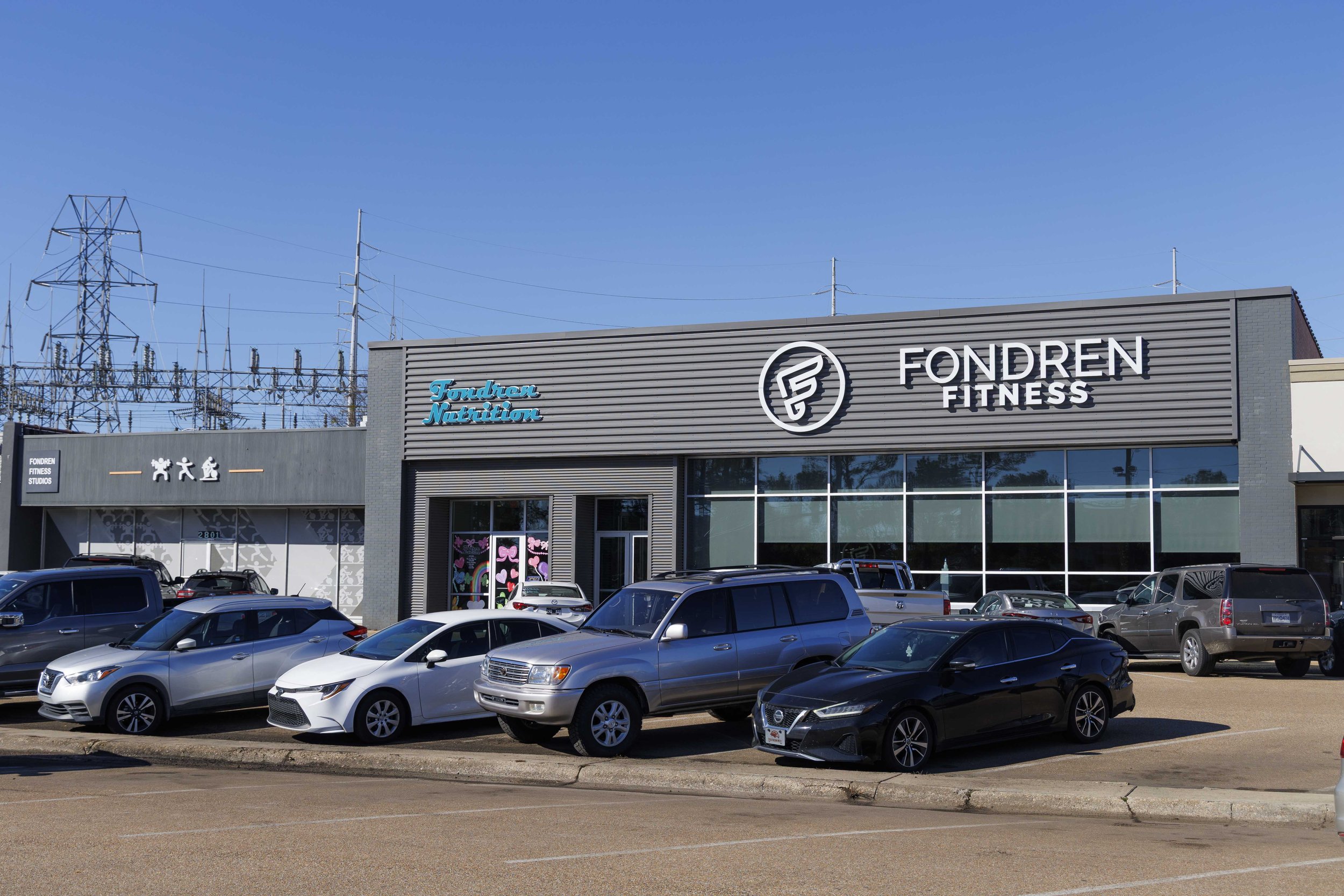 A fitness center named FONDREN FITNESS with a parking lot filled with cars in front. The building has signage for FONDREN Nutrition and various graphics. In the background, there are multiple electrical towers and power lines under a clear blue sky.