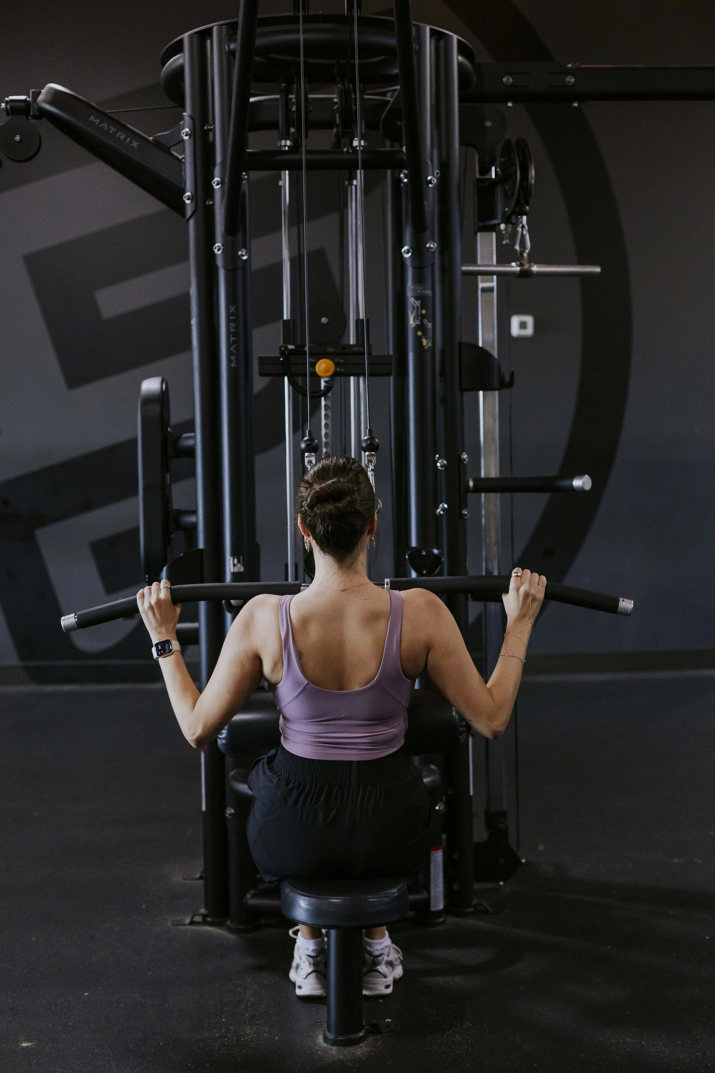 A woman in a purple sports bra and black pants sitting on a black exercise bench, holding a barbell behind her neck, in a dark gym with black walls and gym equipment.