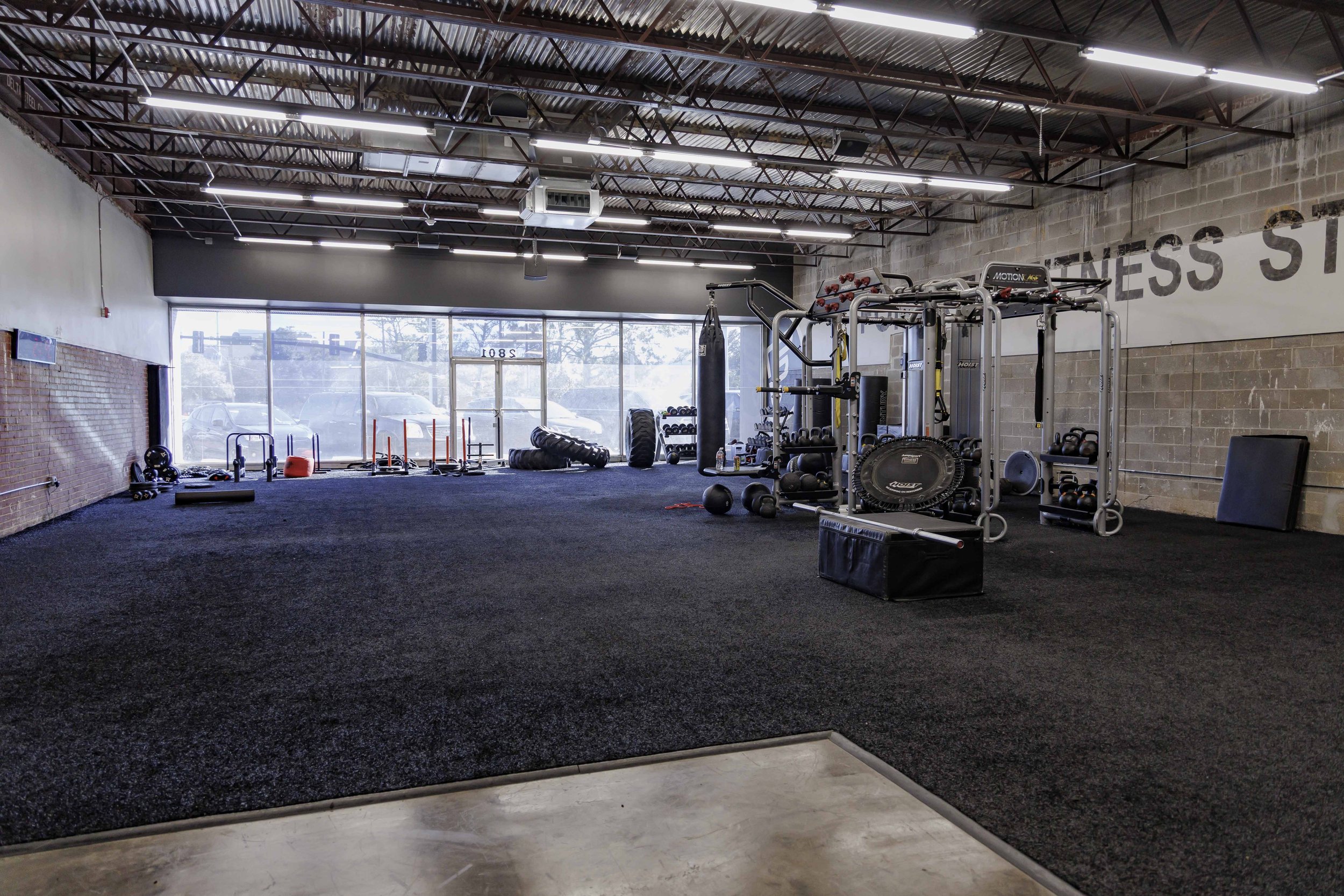 Interior of a modern gym with various workout equipment such as kettlebells, medicine balls, tires, and a power rack, with a large window showing parked cars outside.