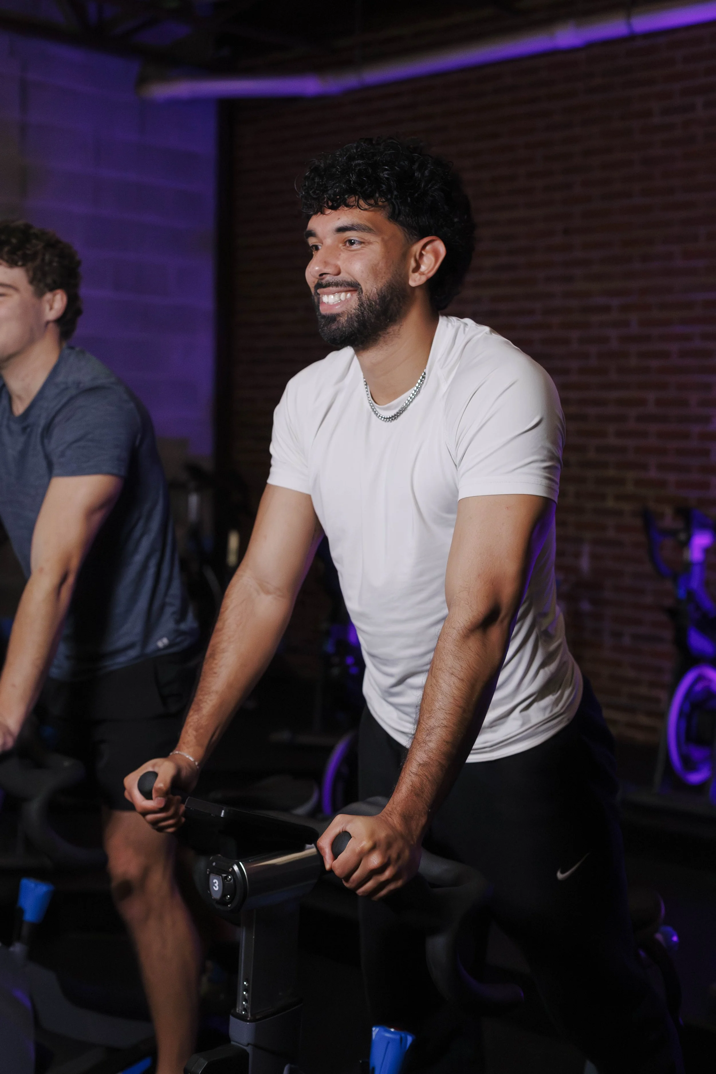 A man with dark curly hair, beard, wearing a white T-shirt and silver chain, is smiling while riding a stationary exercise bike in a gym with a brick wall background and purple lighting.