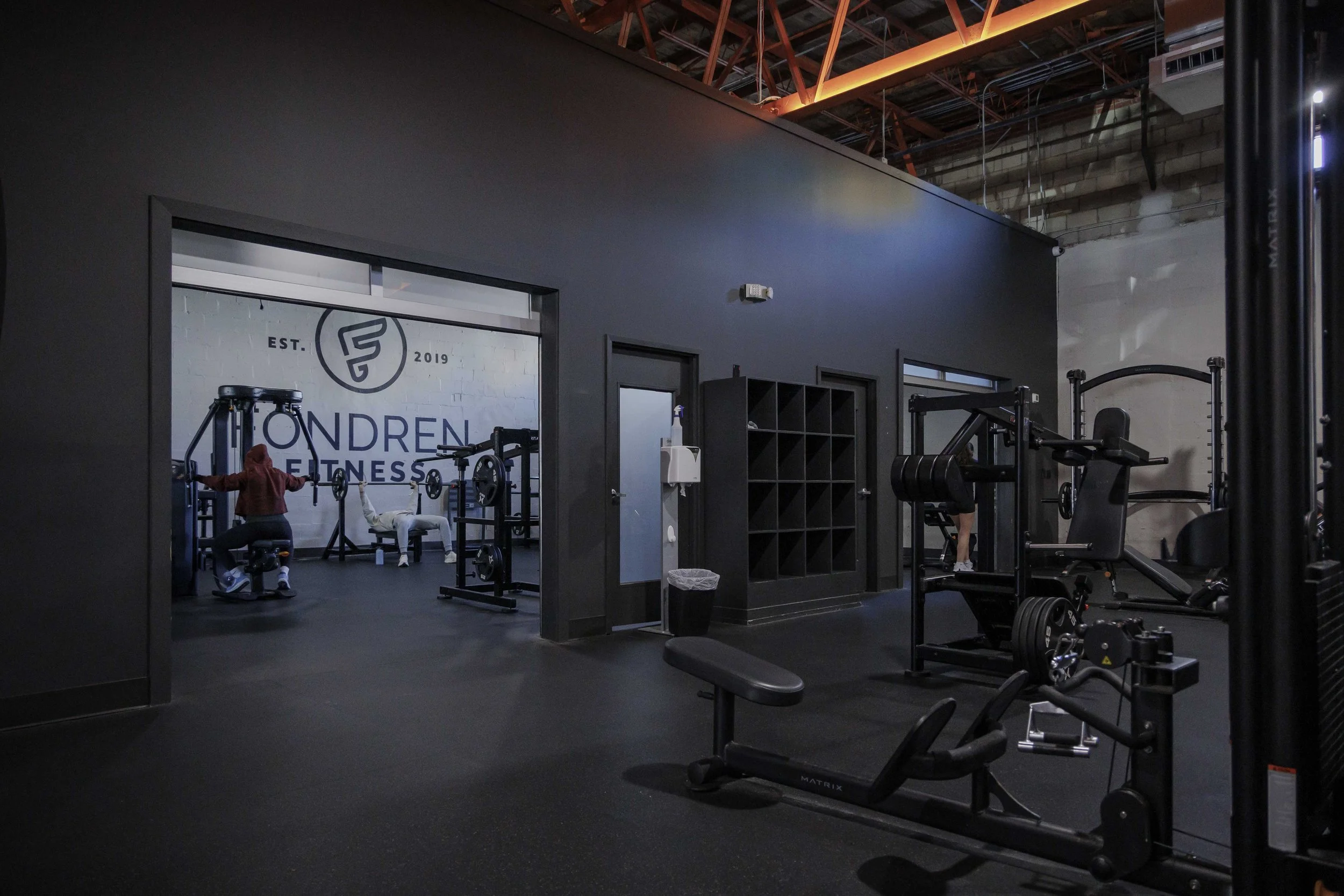 Inside a modern gym with various exercise equipment and a wall with fitness branding. Gym-goers using weights and machines in a well-lit workout area.