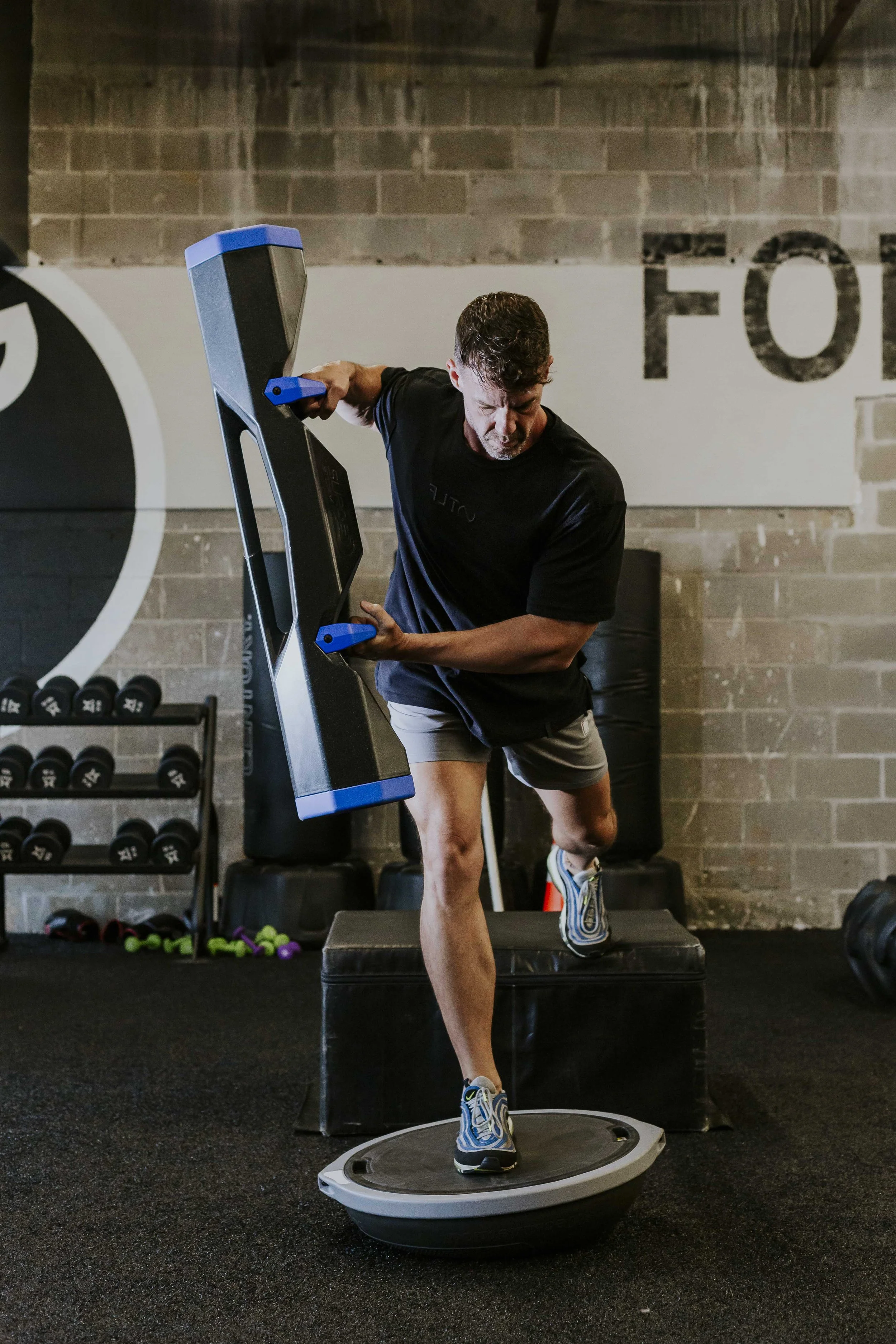 A man performing a workout on a vibration plate machine in a gym, balancing with one foot on the machine and the other foot on a step platform.