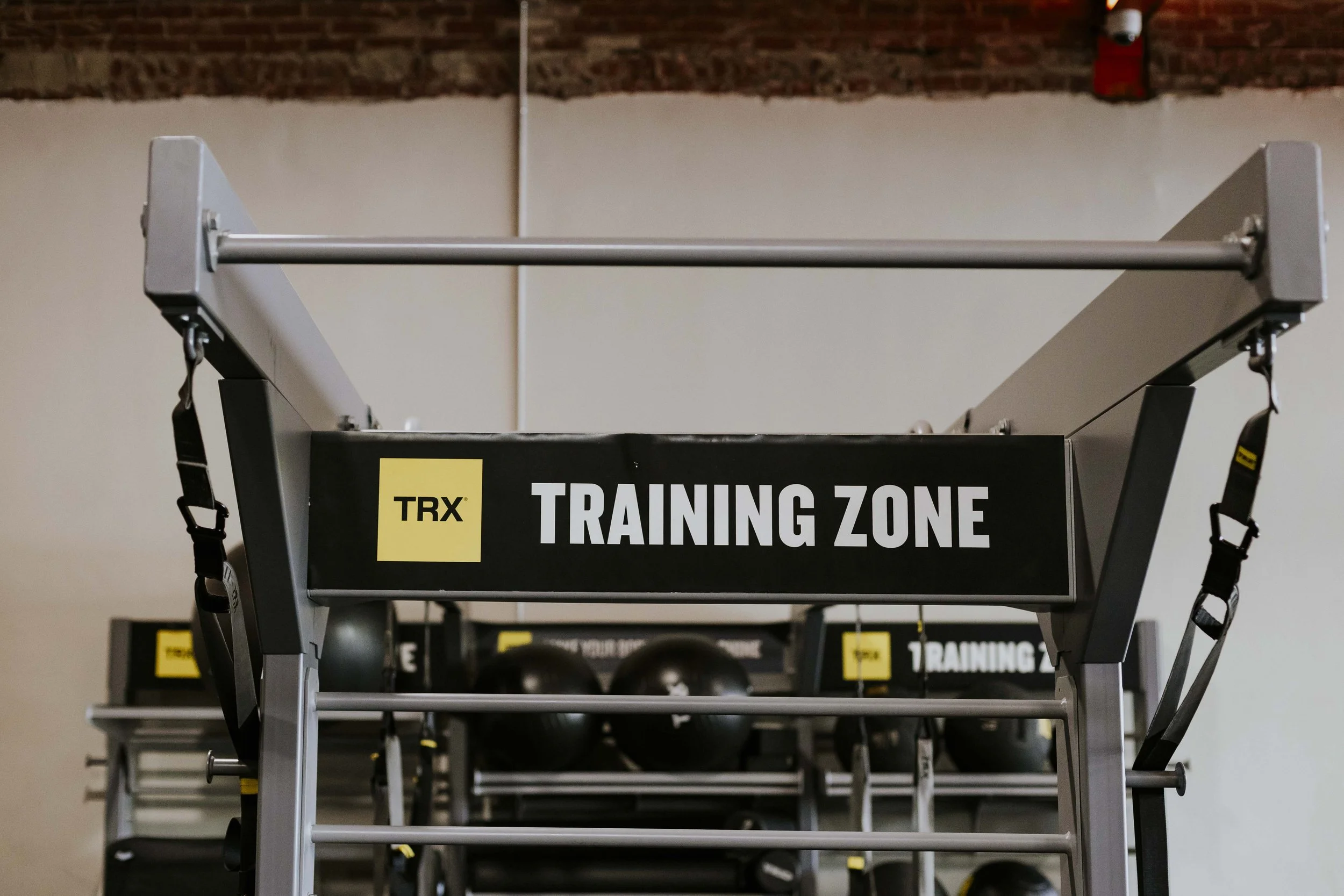 A fitness training station in an indoor gym, with framing equipment labeled 'Training Zone' and black straps hanging from the sides.