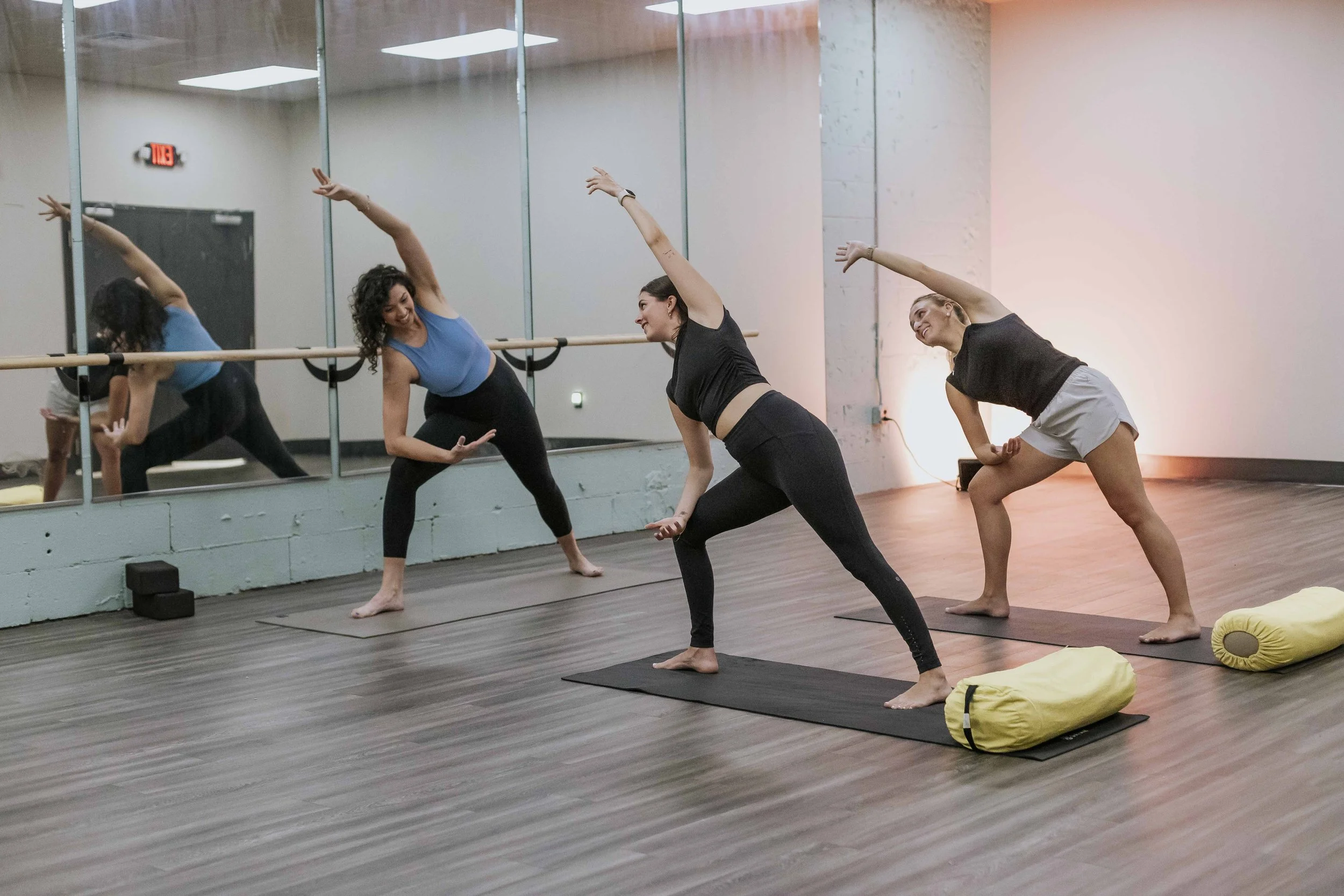 Three women practicing yoga in a studio. They are in a stretching pose with one arm raised and leaning sideways. The studio has a large wall mirror, wooden floors, and yoga mats, with a yellow yoga bolster and black blocks on the floor.