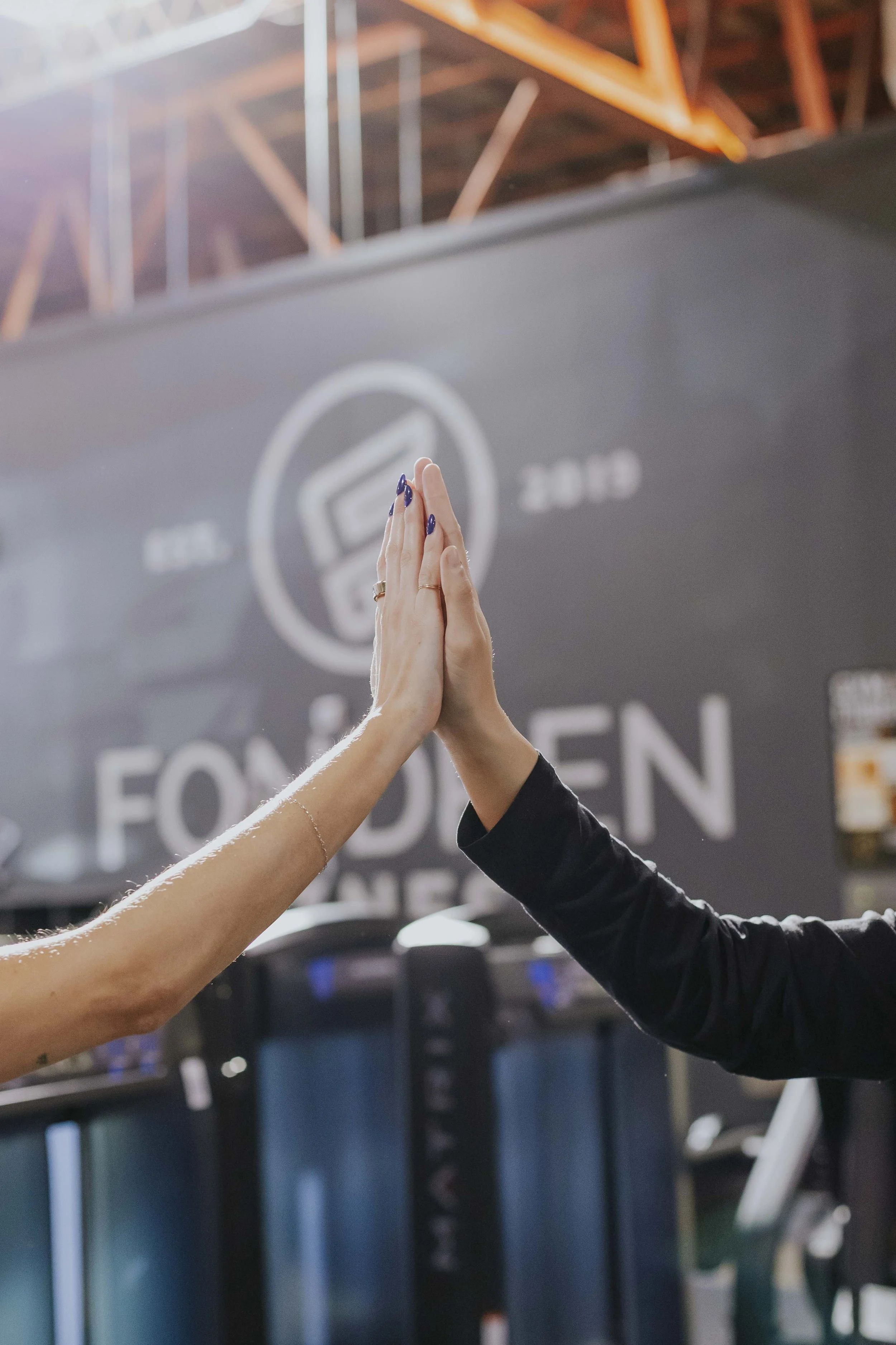 Two people giving each other a high-five with their hands in the center of the frame at a gym or fitness center, with exercise equipment visible in the background.