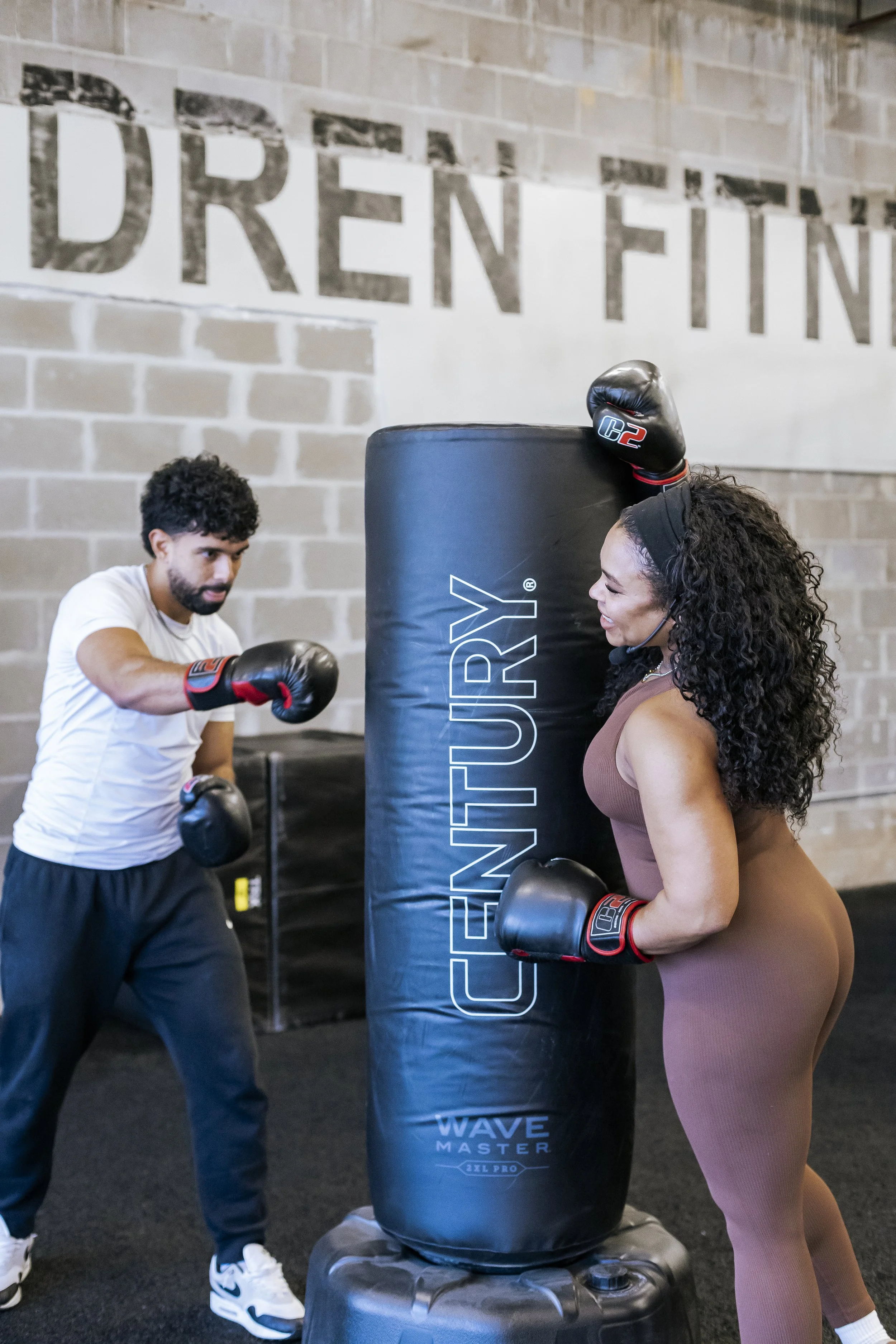 A man and woman practicing boxing with a punching bag at a gym. The man is wearing white T-shirt and black pants, the woman is in brown workout attire. They are both wearing boxing gloves, and the woman is smiling.