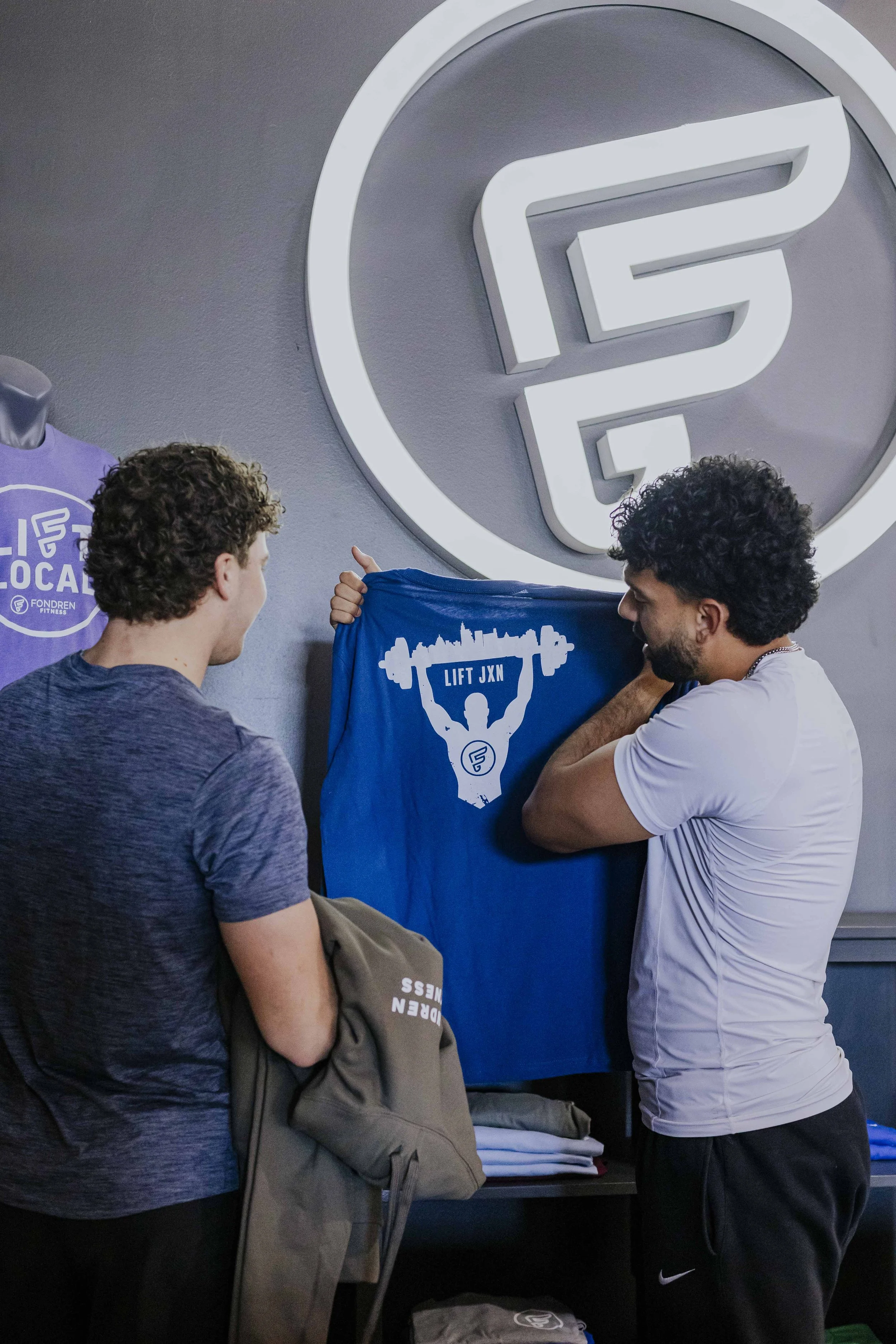Two men are unveiling a blue towel with a white logo that features a silhouette of a person lifting a barbell above their head, with the text "LIFT JXN." They are standing in front of a large illuminated logo of the letter 'G' inside a circle on a gr
