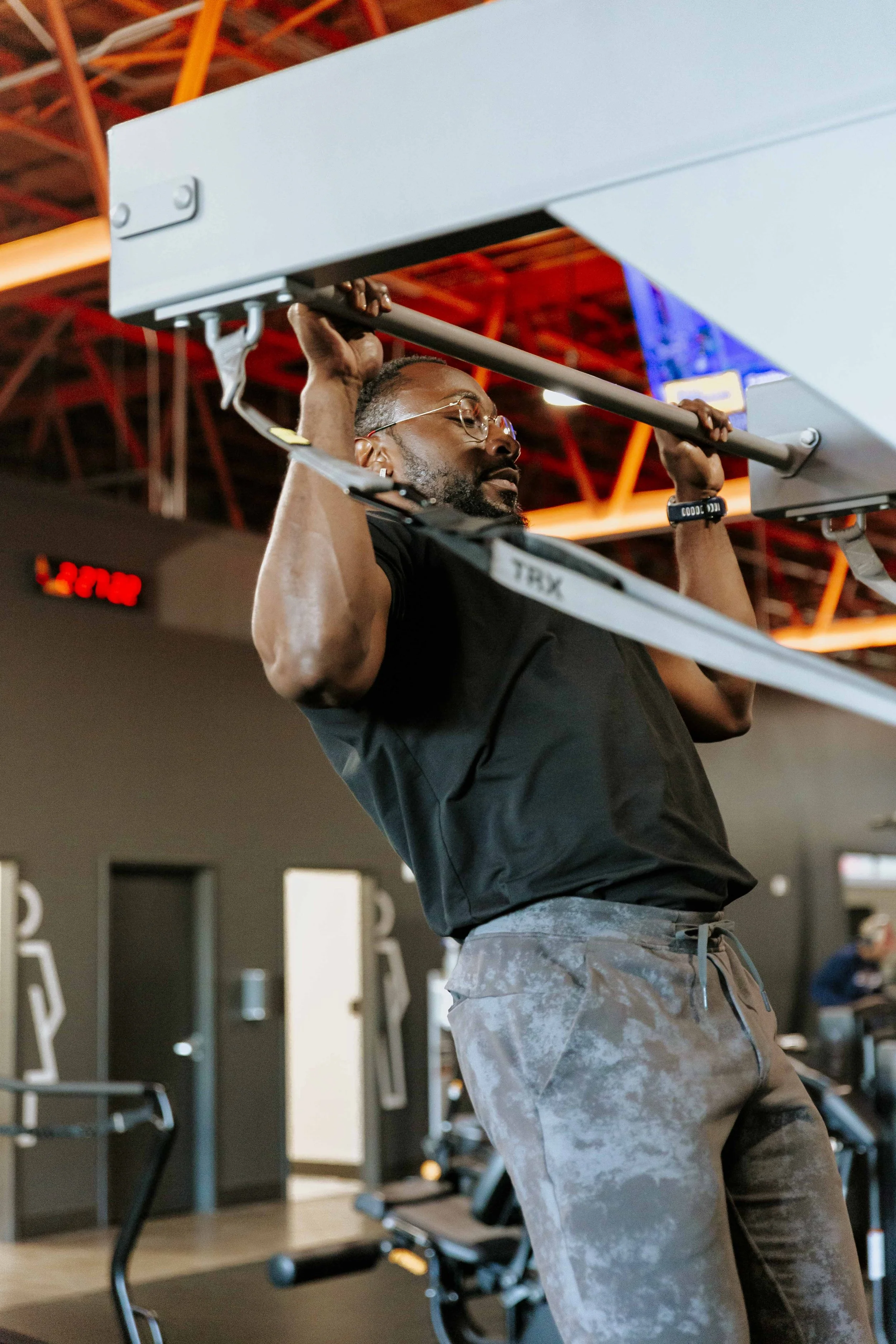 A man with glasses and a beard is doing a pull-up exercise in a gym. He is wearing a black shirt and camouflage shorts, gripping a pull-up bar attached to a machine. The gym has red metal beams and various workout equipment in the background.