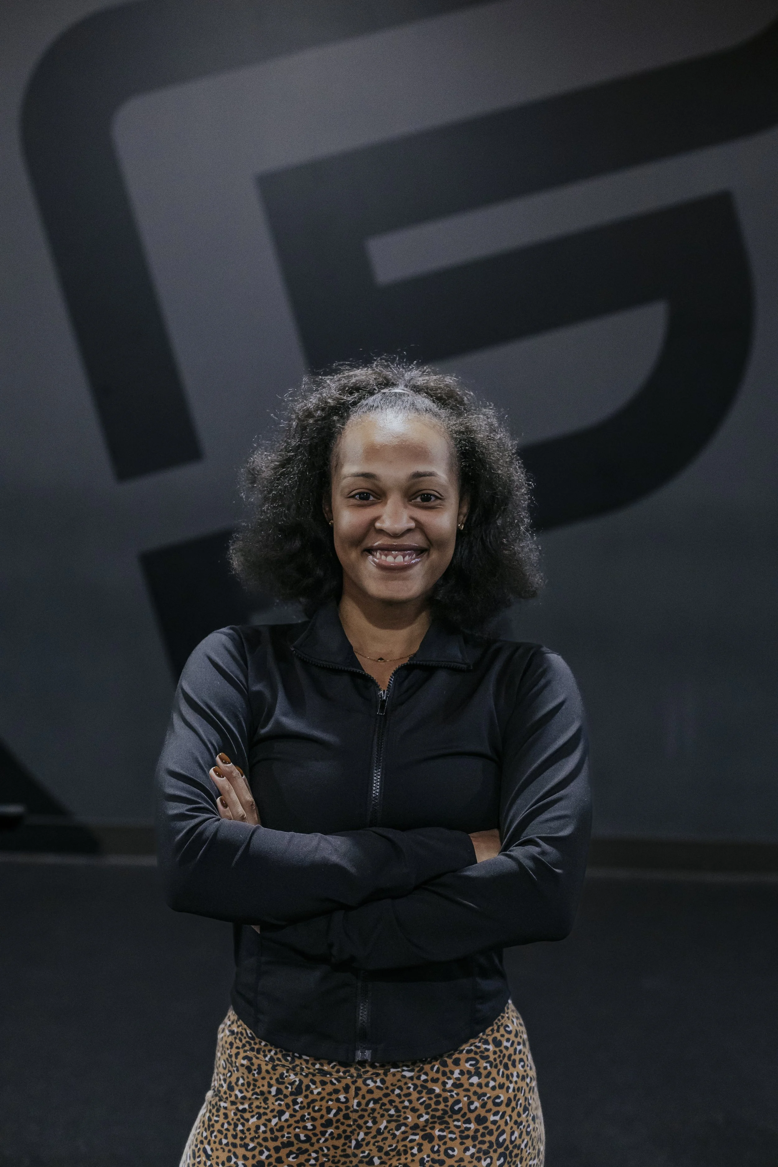 A young woman with curly hair styled in an afro and wearing a black zip-up jacket, standing with arms crossed, smiling in front of a large black wall with a geometric design.