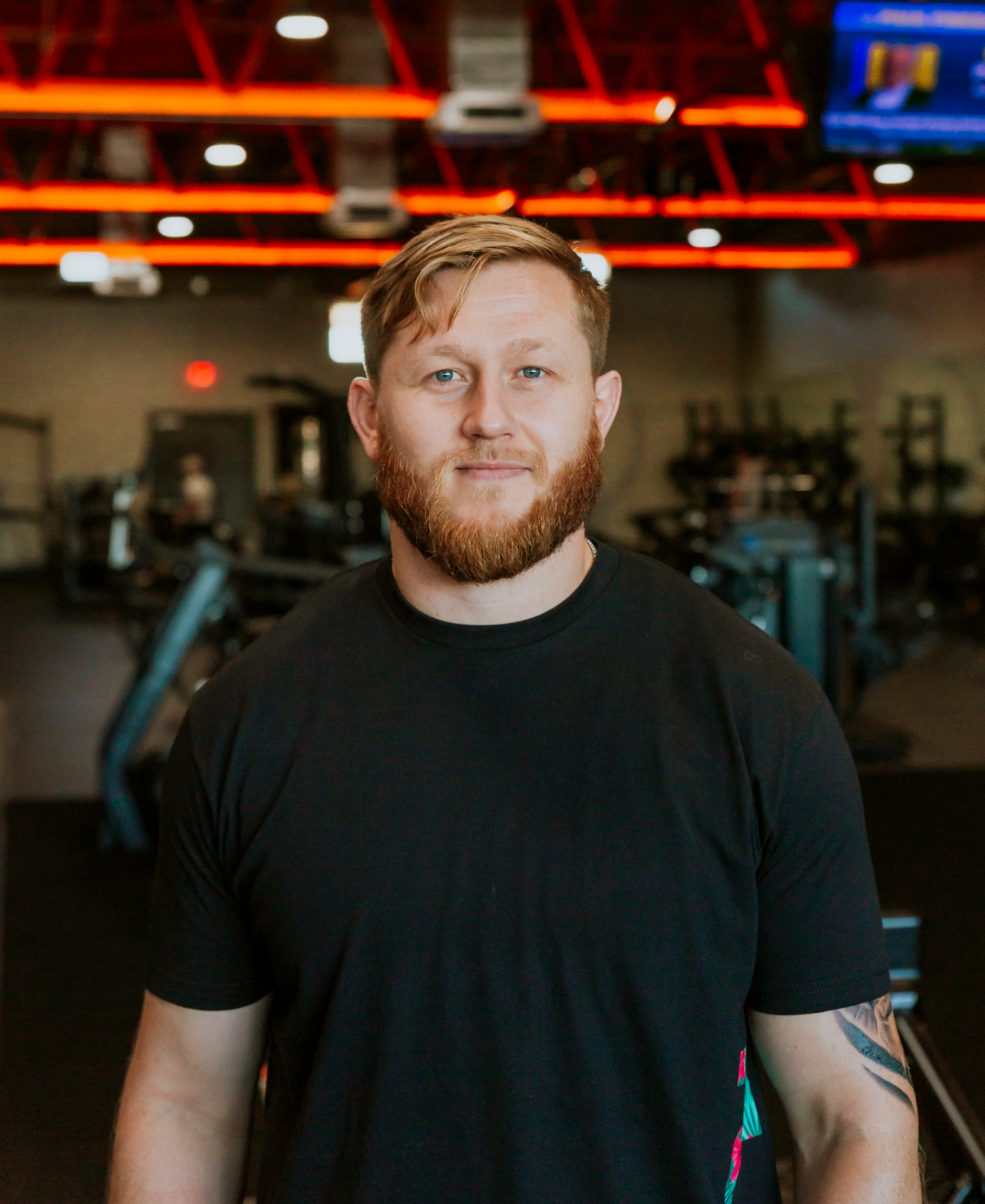 A young man with reddish hair and a full beard wearing a black T-shirt standing in a gym with blurred exercise equipment in the background.