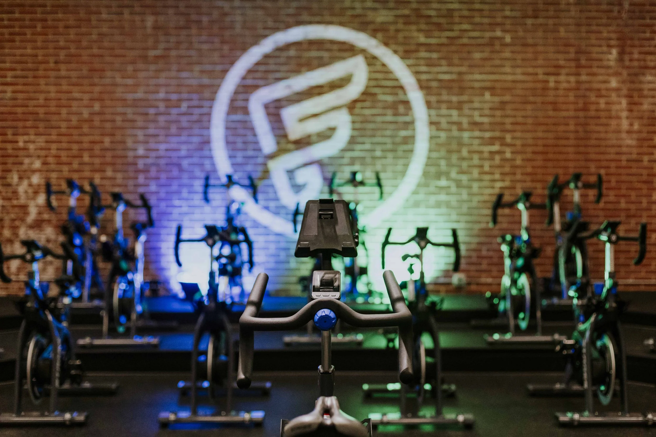 Indoor cycling studio with a brick wall displaying neon lights and artwork, exercise bikes arranged in rows, and a digital handlebar in the foreground.