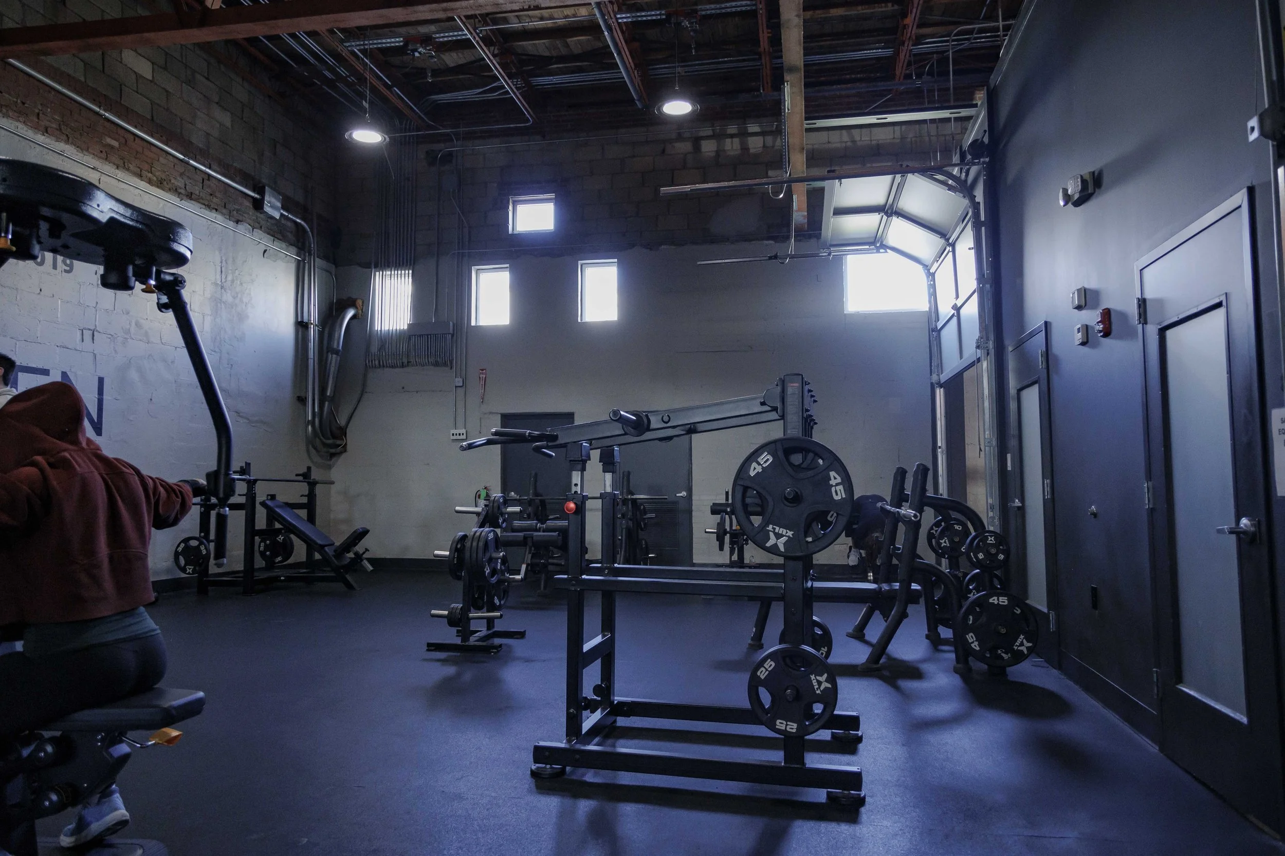 A gym with exercise equipment including weights and a machine, with sunlight coming through high windows, exposed brick wall, and dark floors.