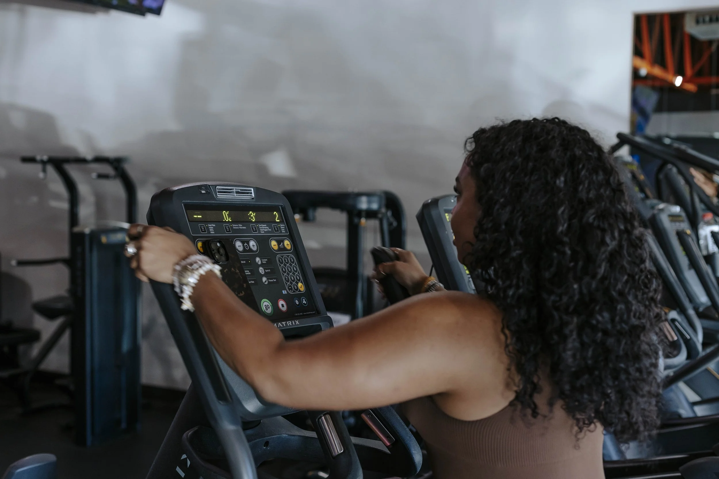 A woman with curly black hair riding or training on a stationary exercise bike in an indoor gym or fitness studio.