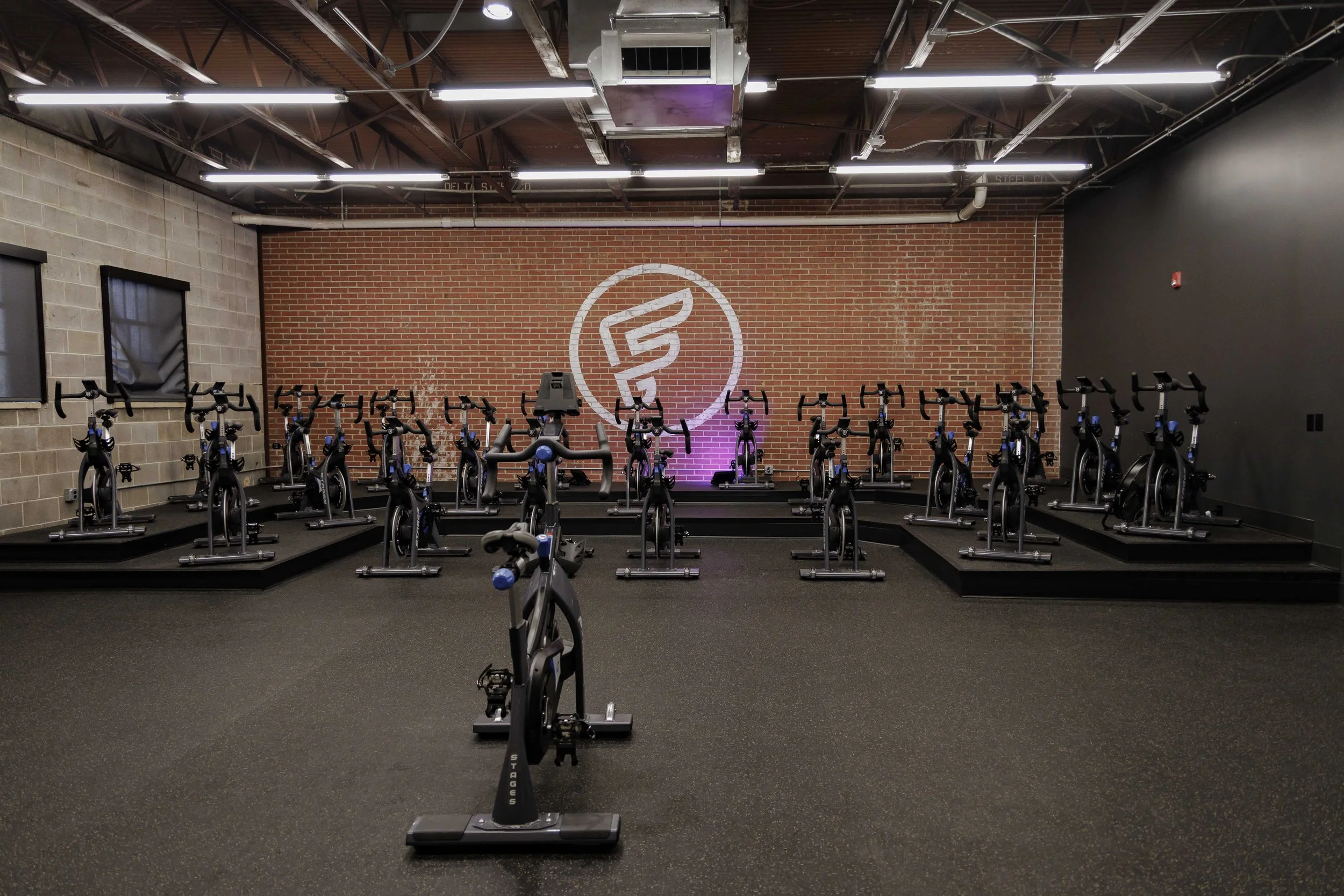 Indoor cycling studio with multiple stationary bikes arranged in rows on black platforms, a large red brick wall with a logo, exposed ceiling with lighting and ventilation, and a dark-colored wall.