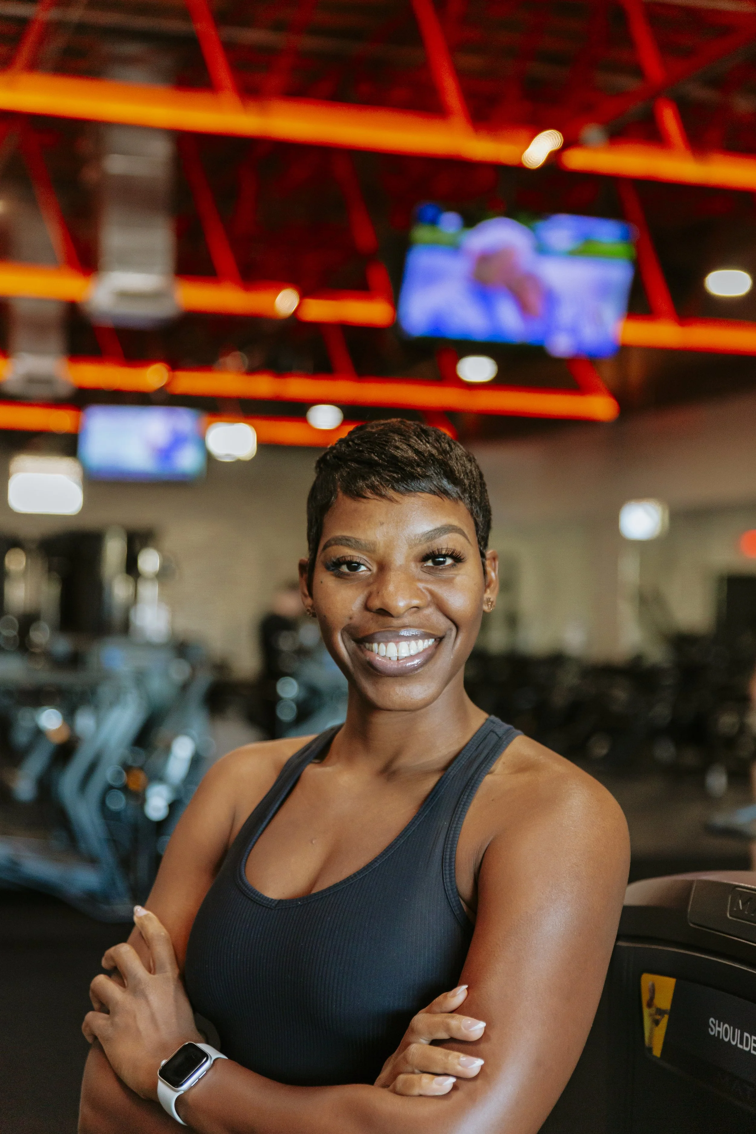 A young woman with short black hair smiling at the gym, wearing a black sports tank top and a fitness tracker on her wrist, with gym equipment and exercise bikes in the background.