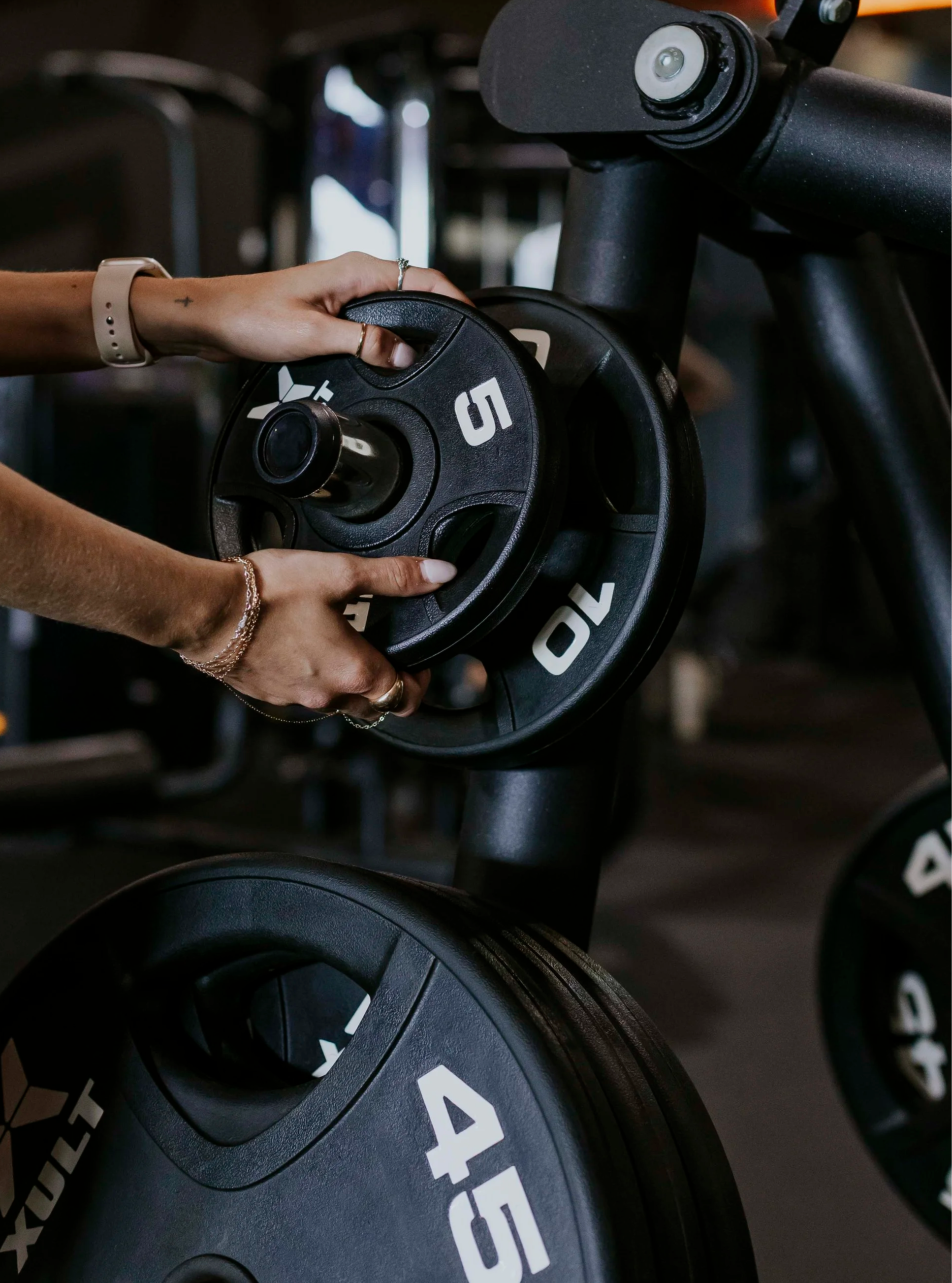 A person with a small tattoo on their wrist and wearing rings on their fingers is placing a black weight plate onto a gym wheel or barbell. The gym equipment displays the word 'FITS' in white letters.