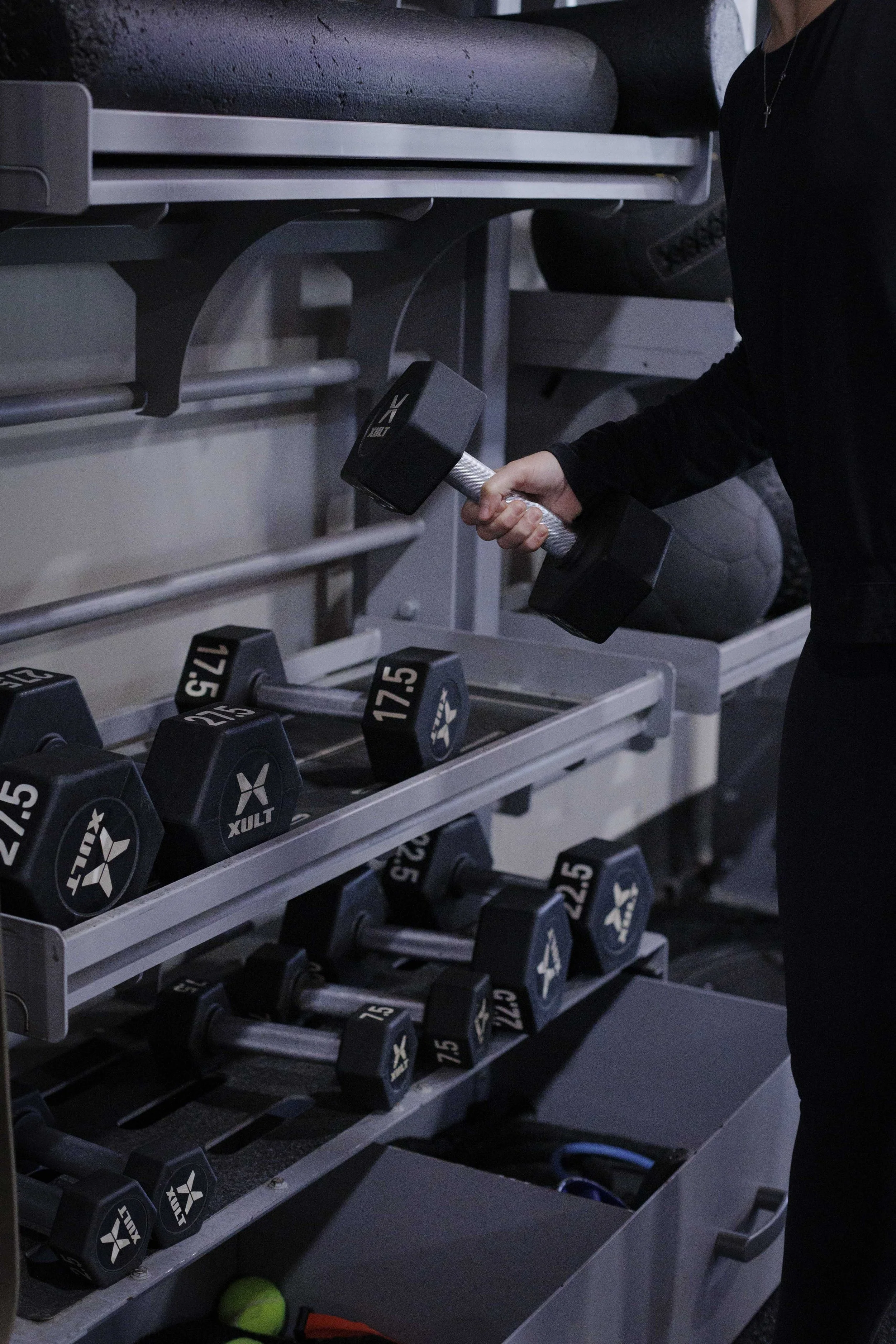 Person lifting a black dumbbell in a gym with various other weights on a rack.